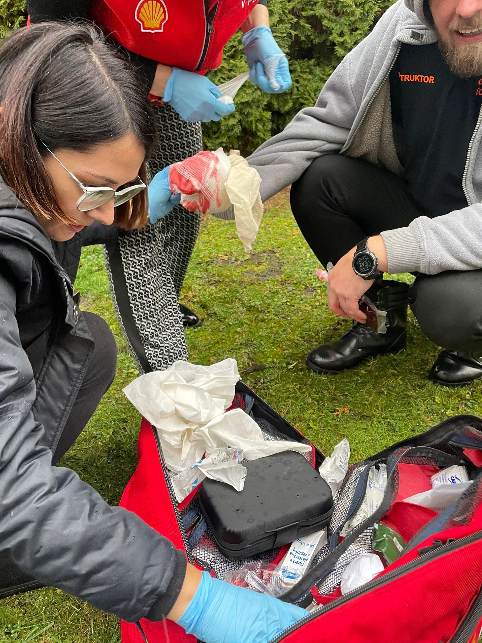 Opatrywanie zakrwawionej ręki na trawie. Osoby w rękawiczkach, apteczka pierwszej pomocy, opatrunki i środki dezynfekcyjne.