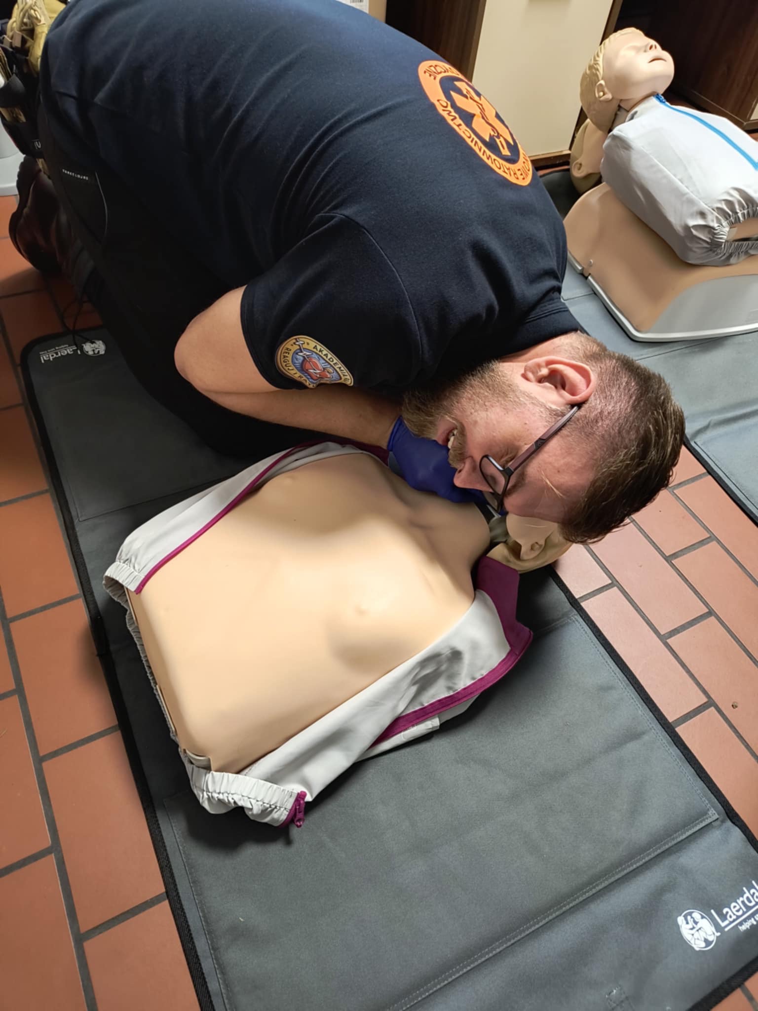 Instruktor w okularach, w maseczce i koszulce z logo ratownictwa, demonstruje usta-usta na manekinie podczas zajęć z pierwszej pomocy. W tle drugi manekin.