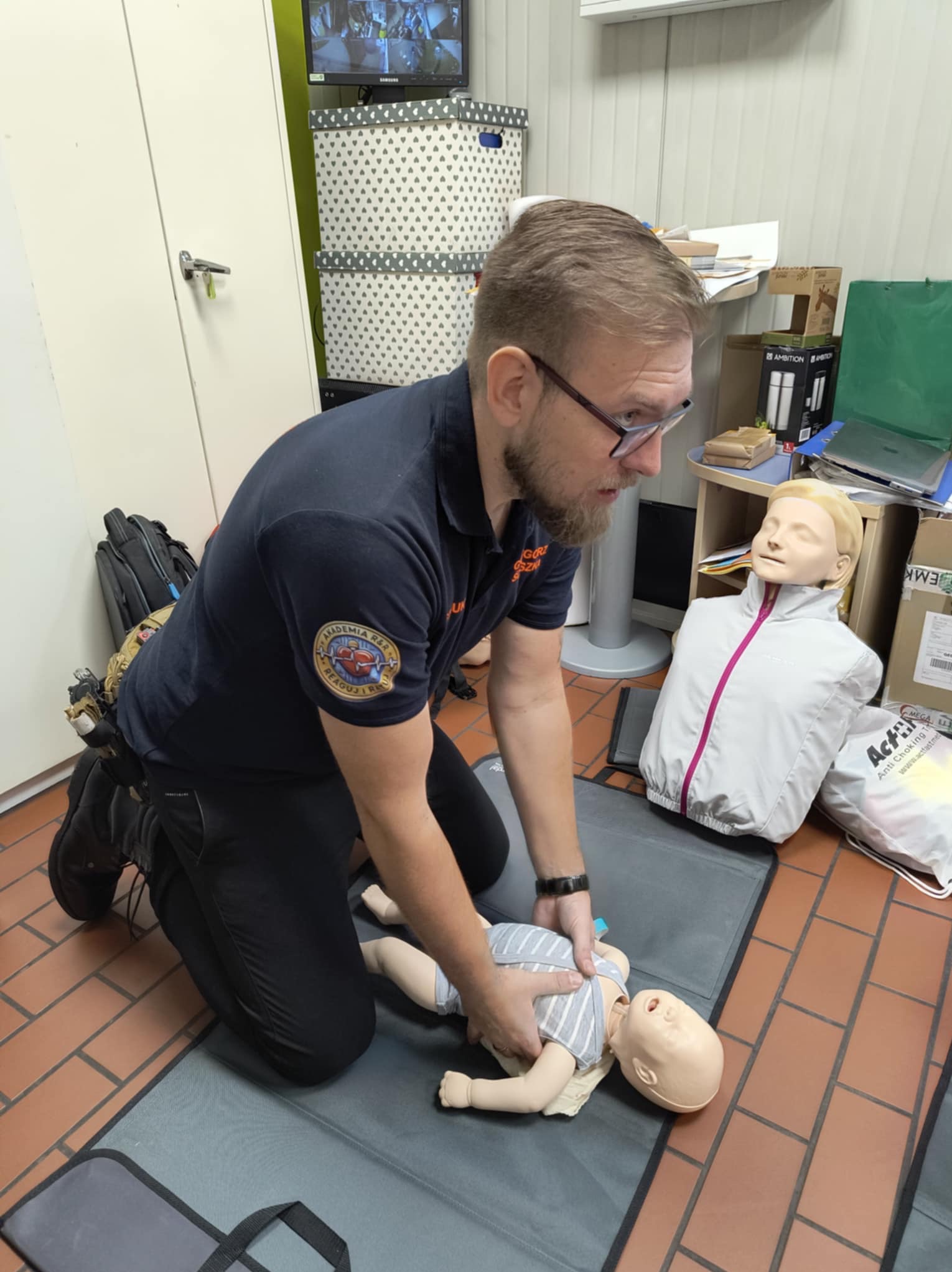 Instruktor z brodą i okularami w Bielsku-Białej demonstruje na manekinie niemowlęcia technikę pierwszej pomocy, obok leży manekin osoby dorosłej, w tle widać szafki i monitor z obrazem z kamer...