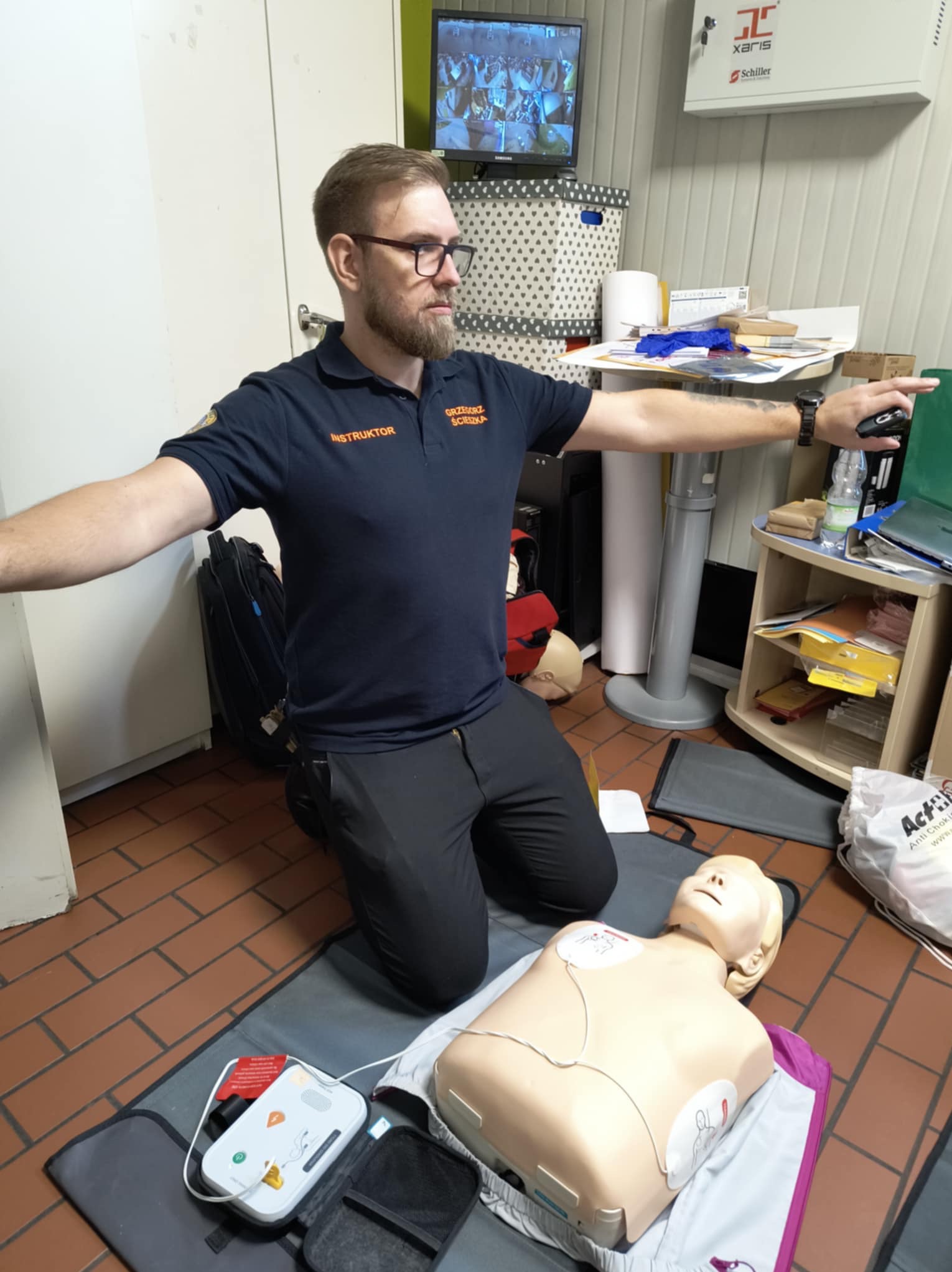 Instruktor w okularach, w granatowej koszulce polo z napisem 'Instruktor' i nazwiskiem, demonstruje użycie AED na manekinie podczas zajęć. W tle monitor z obrazem z kamer monitoringu.