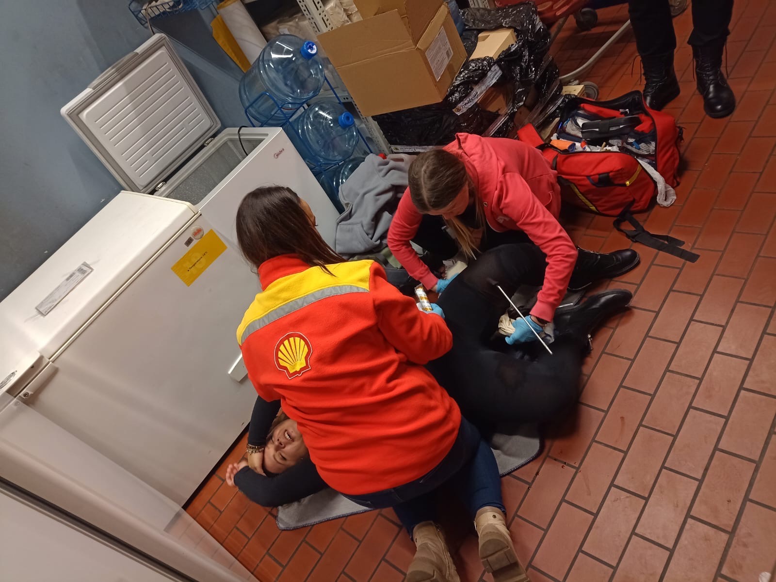 Dwie kobiety w odzieży roboczej udzielają pierwszej pomocy osobie leżącej na podłodze w pomieszczeniu z lodówkami i kartonami, jedna z kobiet ma na ubraniu logo Shell.