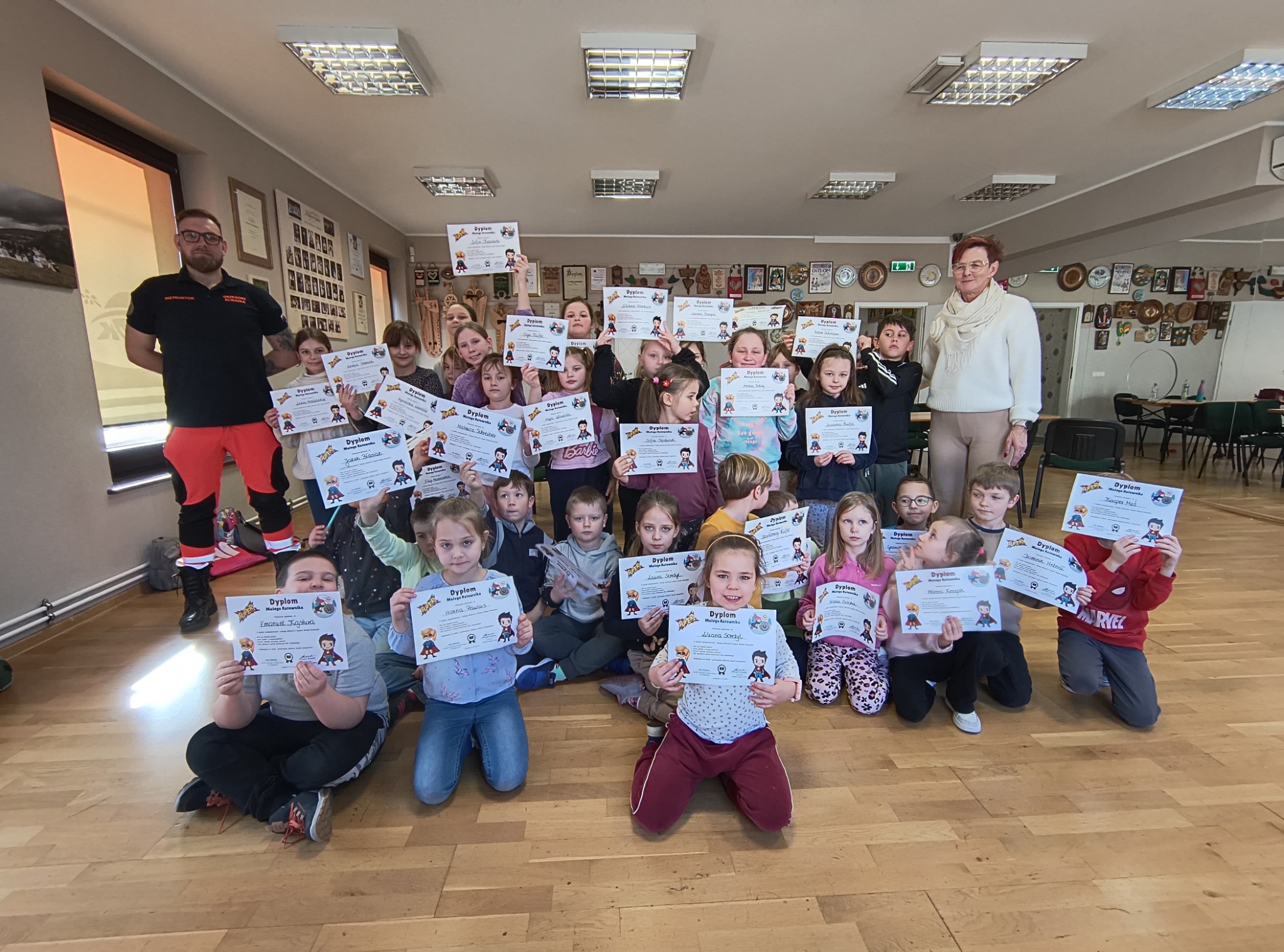 Grupa dzieci trzymająca dyplomy Małego Ratownika na tle sali z instruktorem w kombinezonie ratowniczym i kobietą w swetrze.