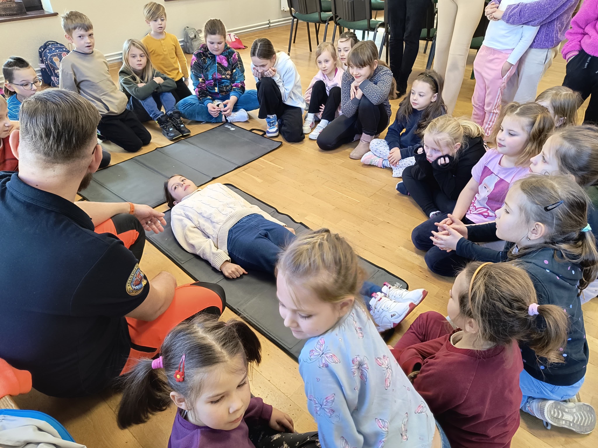 Grupa dzieci siedzi na podłodze w kręgu, obserwując instruktora demonstrującego techniki ratownicze na manekinie leżącym na czarnej macie. Instruktor w czarnej koszuli z naszywką na ramieniu,...