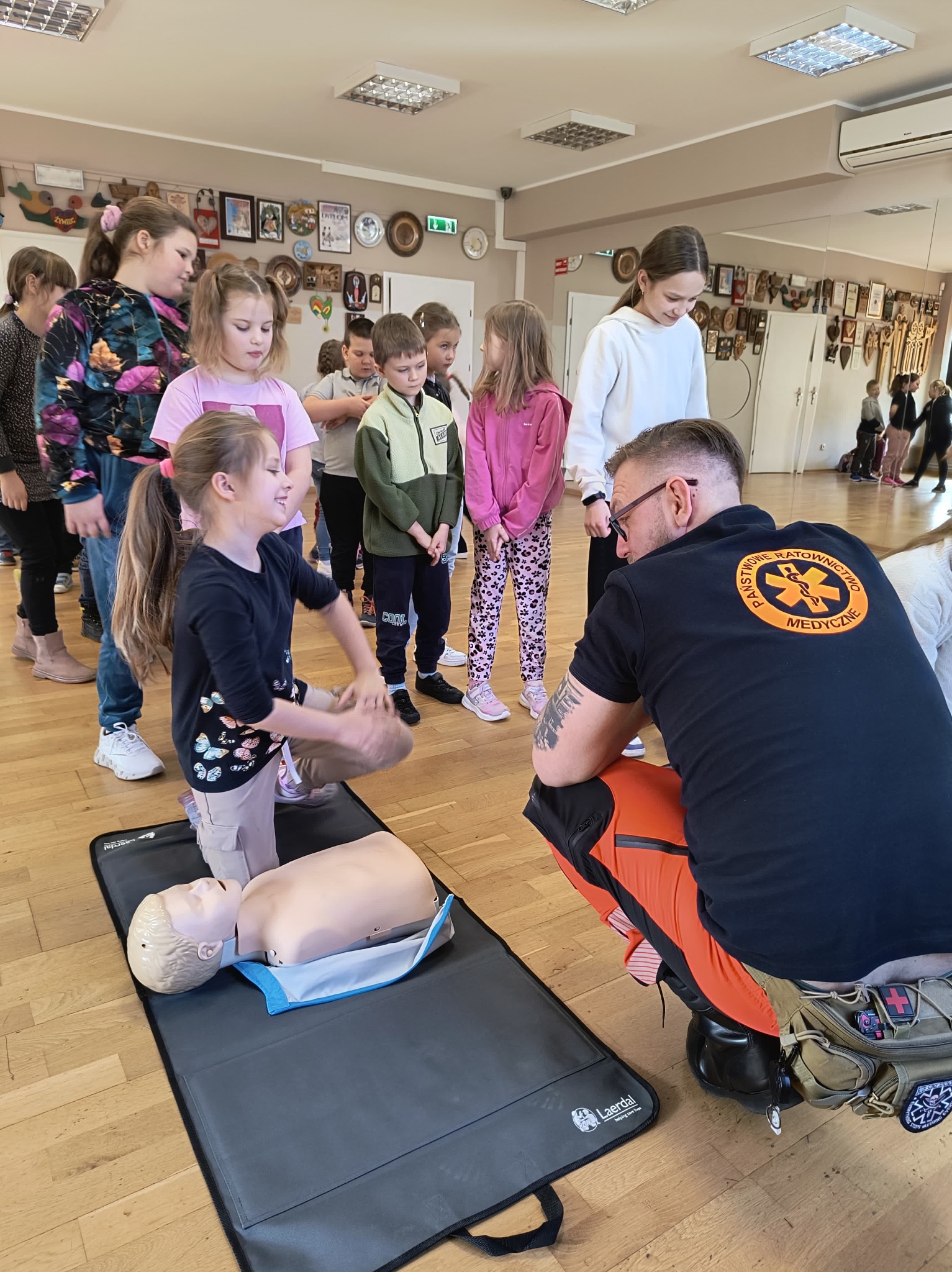 Dziewczynka ćwiczy resuscytację krążeniowo-oddechową na fantomie pod okiem instruktora, grupa dzieci obserwuje, w tle sala z lustrami i dekoracjami.