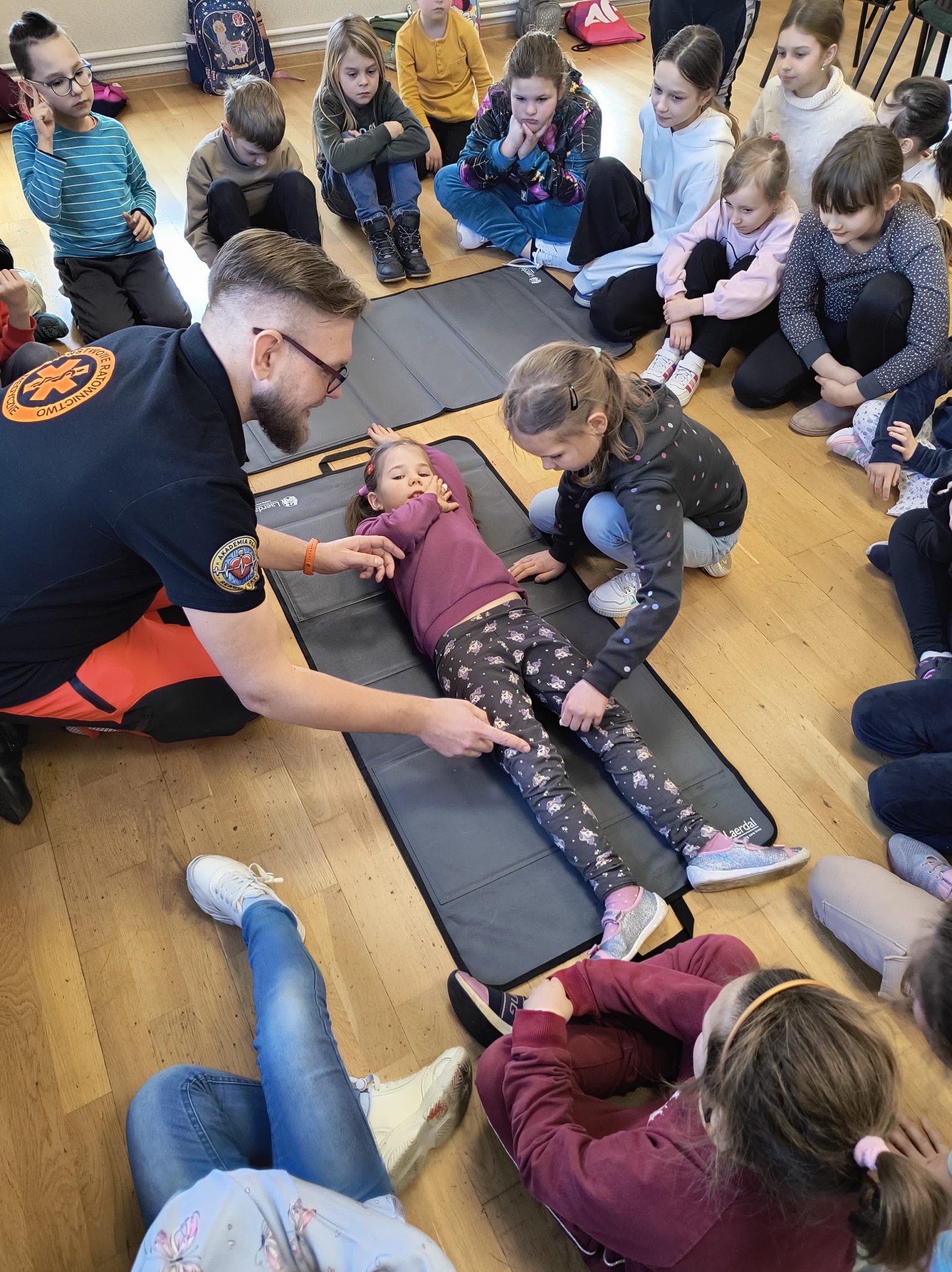Instruktor w czarnej koszulce z logo ratownictwa medycznego demonstruje na dziewczynce leżącej na szarej macie, jak udzielić pierwszej pomocy, w otoczeniu grupy dzieci siedzących na podłodze.