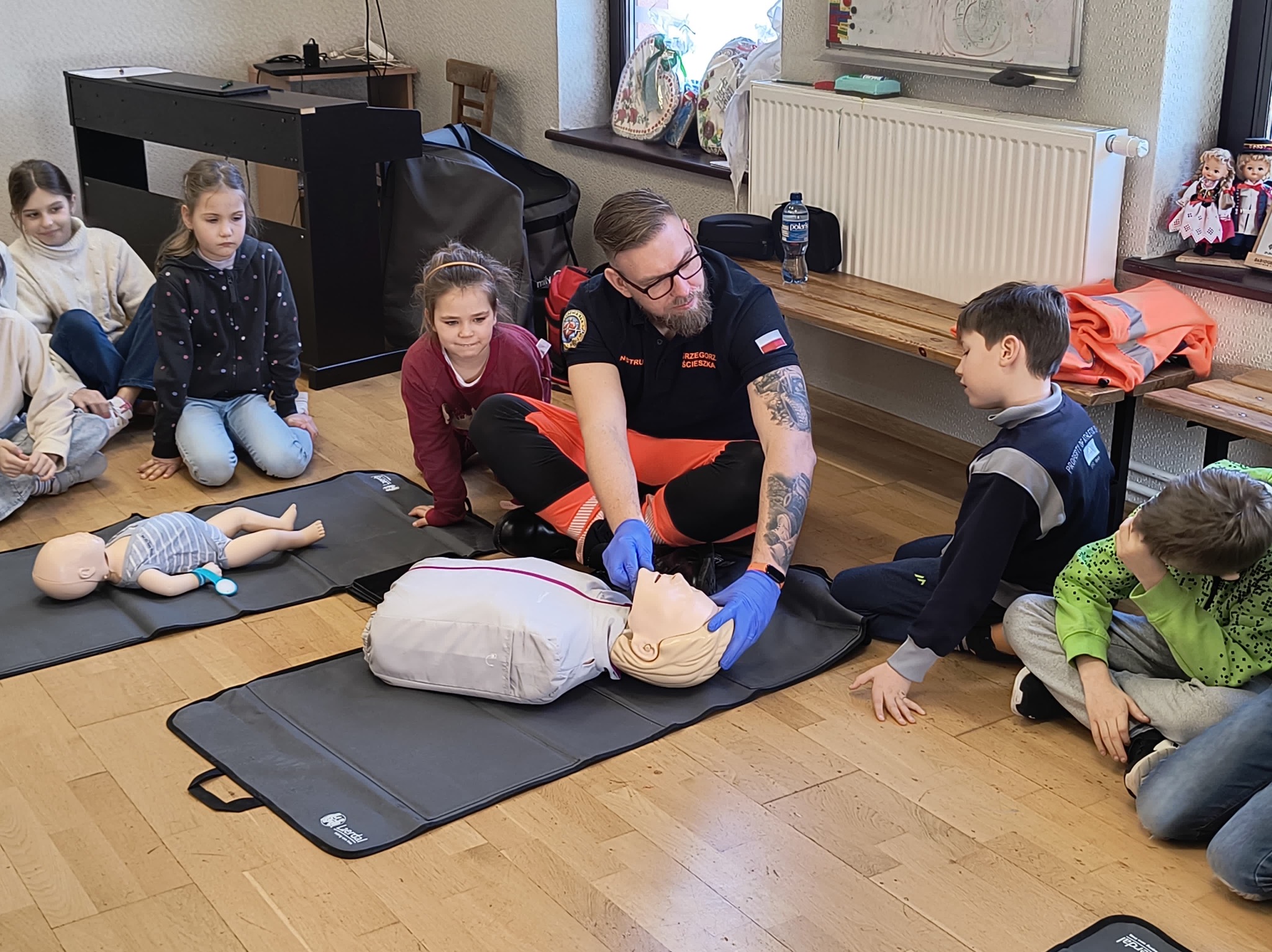 Instruktor w okularach, z tatuażami na ramionach, w niebieskich rękawiczkach demonstruje na manekinie resuscytację usta-usta, otoczony przez grupę dzieci siedzących na podłodze w sali.