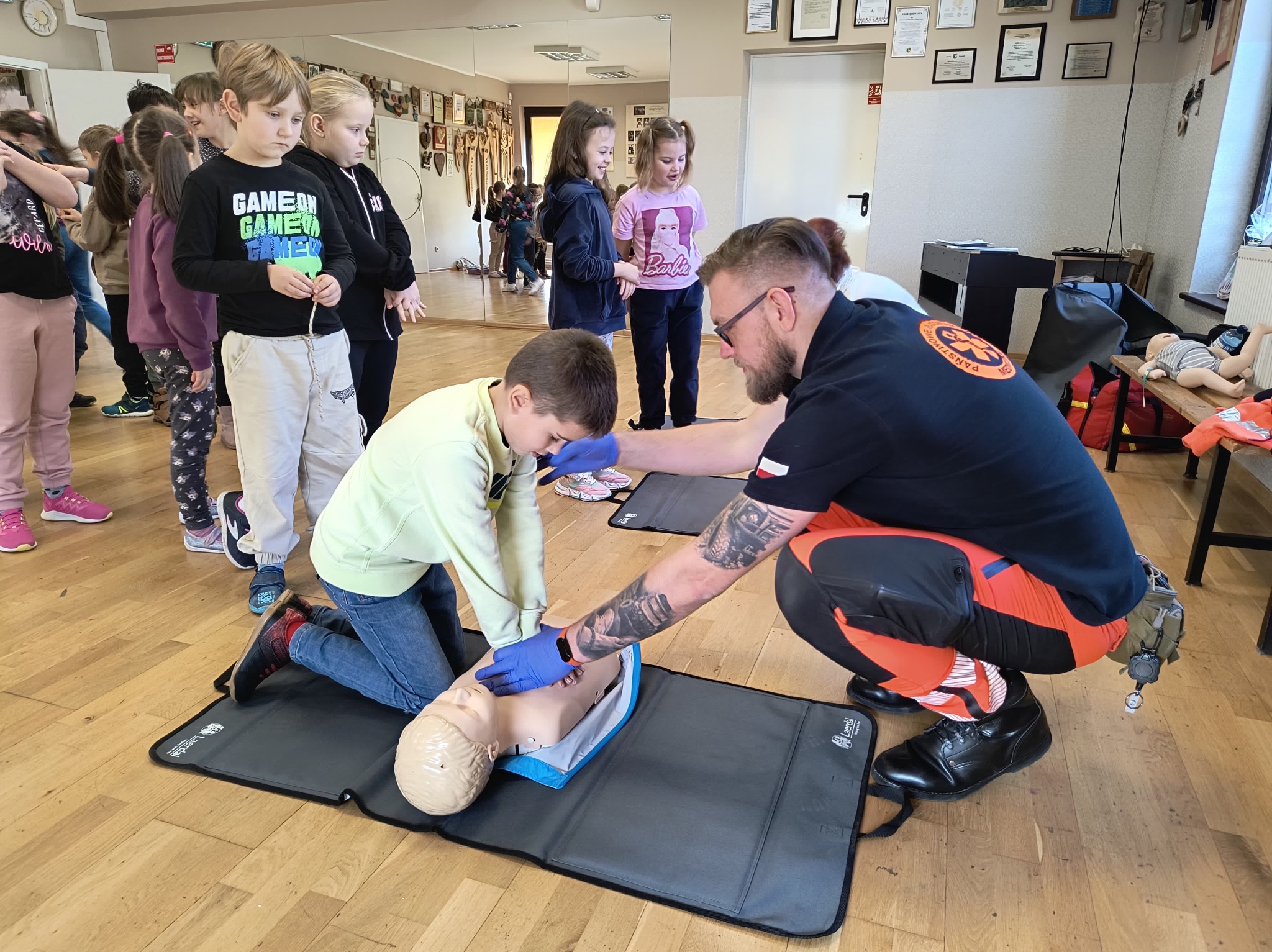 Chłopiec w zielonej bluzie ćwiczy resuscytację krążeniowo-oddechową na manekinie pod nadzorem instruktora w pomarańczowo-czarnym kombinezonie, grupa dzieci obserwuje w tle, na ścianie wiszą dyplomy.