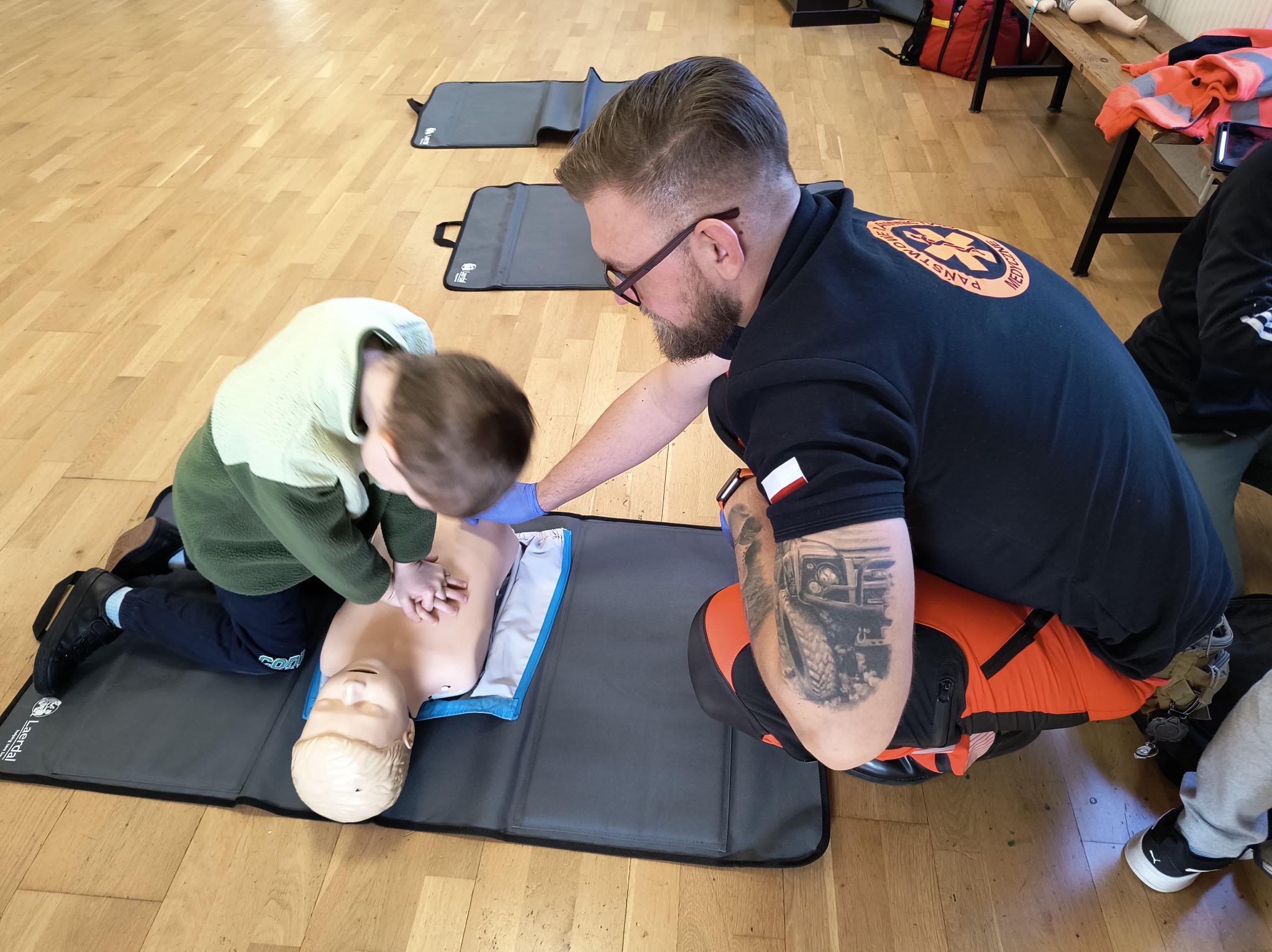 Instruktor w okularach demonstruje na manekinie resuscytację krążeniowo-oddechową dziecku podczas zajęć, widoczny tatuaż na ramieniu instruktora.