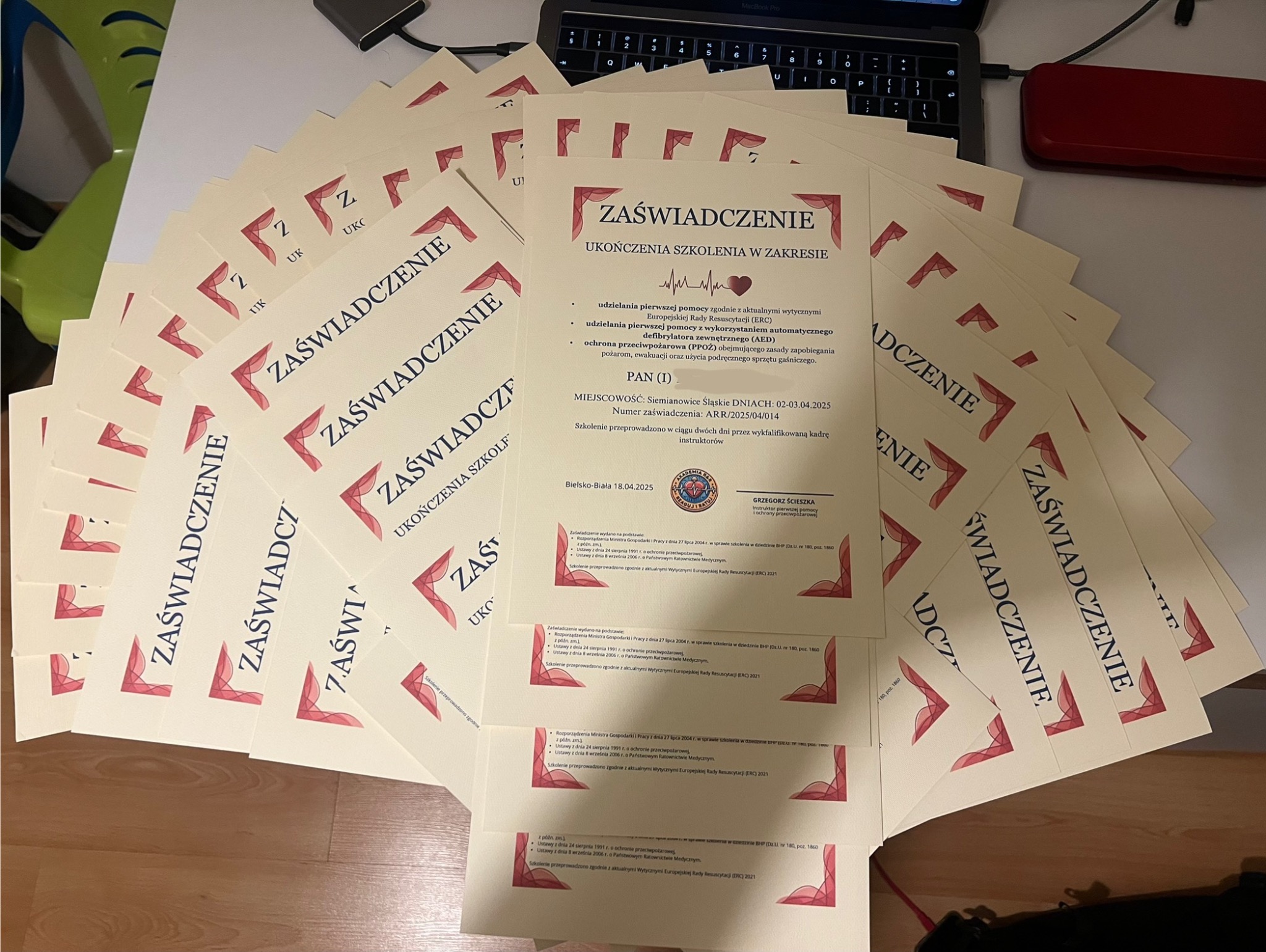 Stos zaświadczeń ukończenia szkolenia z zakresu udzielania pierwszej pomocy, z datami ważności do 2025 roku, leżących na biurku obok laptopa. Dokumenty z logo i danymi instruktora.