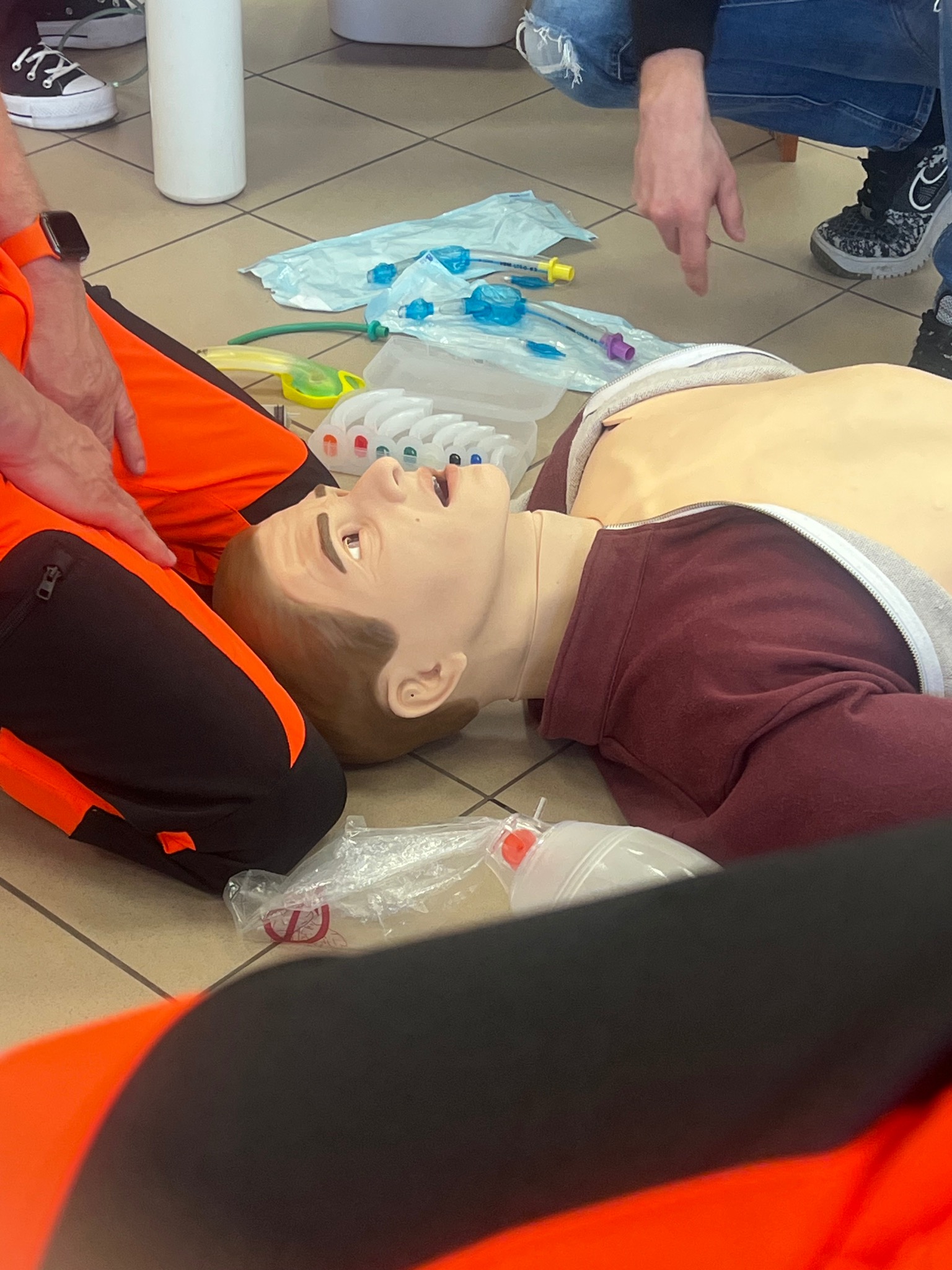 Manekin do nauki pierwszej pomocy, otoczony przez osoby w strojach ratowniczych i sprzęt medyczny, podczas szkolenia z zakresu resuscytacji krążeniowo-oddechowej.