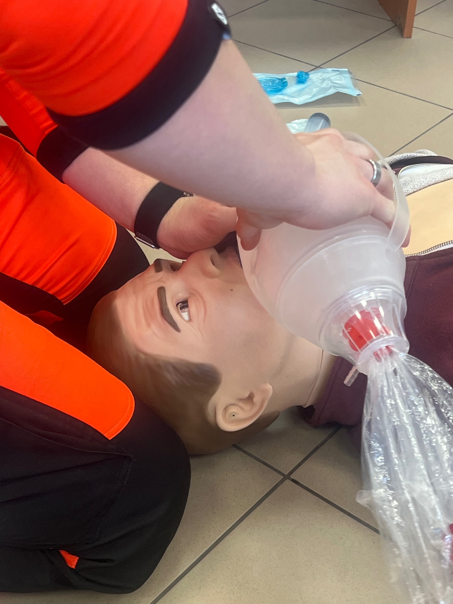 Instruktor w pomarańczowym stroju demonstruje resuscytację usta-usta na fantomie z użyciem worka samorozprężalnego. Widoczny ring na palcu instruktora i płytki podłogowe.