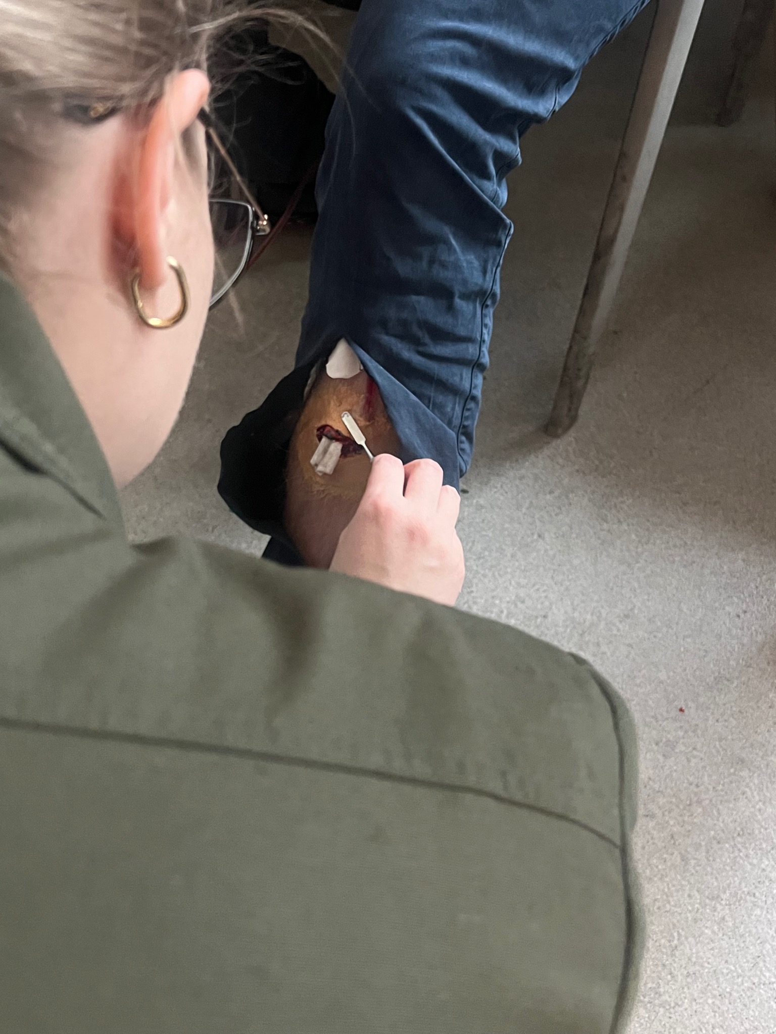 Instruktor demonstruje opatrywanie rany na nodze pozoranta podczas kursu pierwszej pomocy. Widoczne narzędzie i bandaże. Uczestnik z bliska, widoczny fragment ucha i okulary.