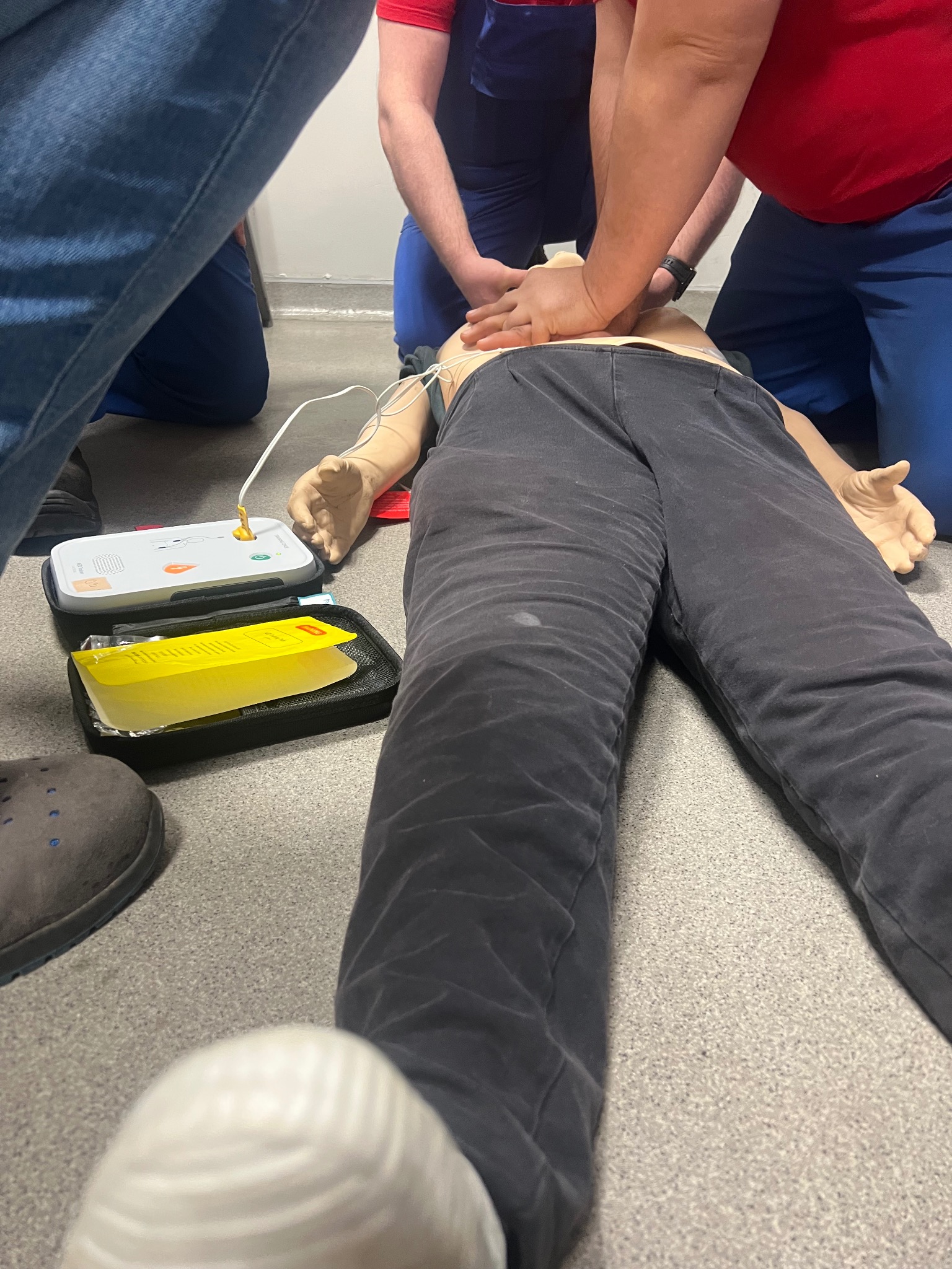 Szkolenie z pierwszej pomocy: resuscytacja krążeniowo-oddechowa na manekinie, widoczny defibrylator i instruktorzy. Ujęcie z góry, detale wyposażenia.