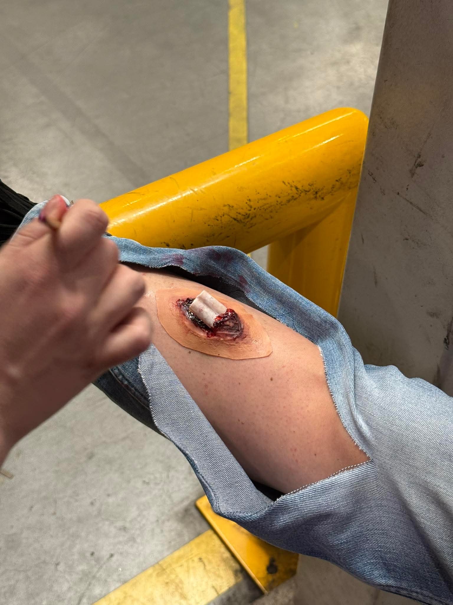 Symulacja rany na nodze, zabezpieczona plastrem z gazą, obok dłoń osoby aplikującej preparat. Scena z kursu pierwszej pomocy, na tle żółtej barierki i betonowej podłogi.