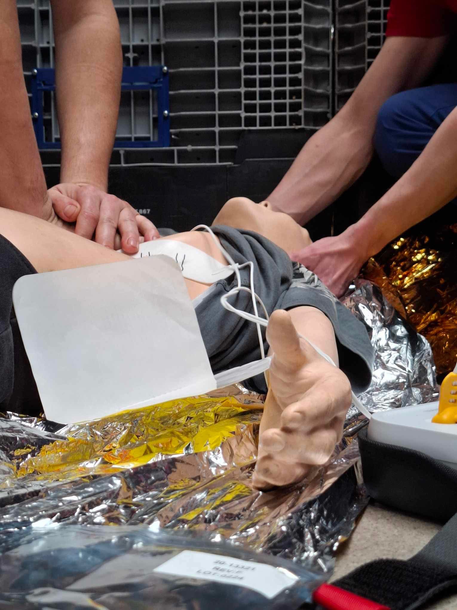 Kurs pierwszej pomocy: Instruktorzy ćwiczą resuscytację na manekinie z widocznym zabezpieczeniem, podkładem termicznym i sprzętem medycznym.