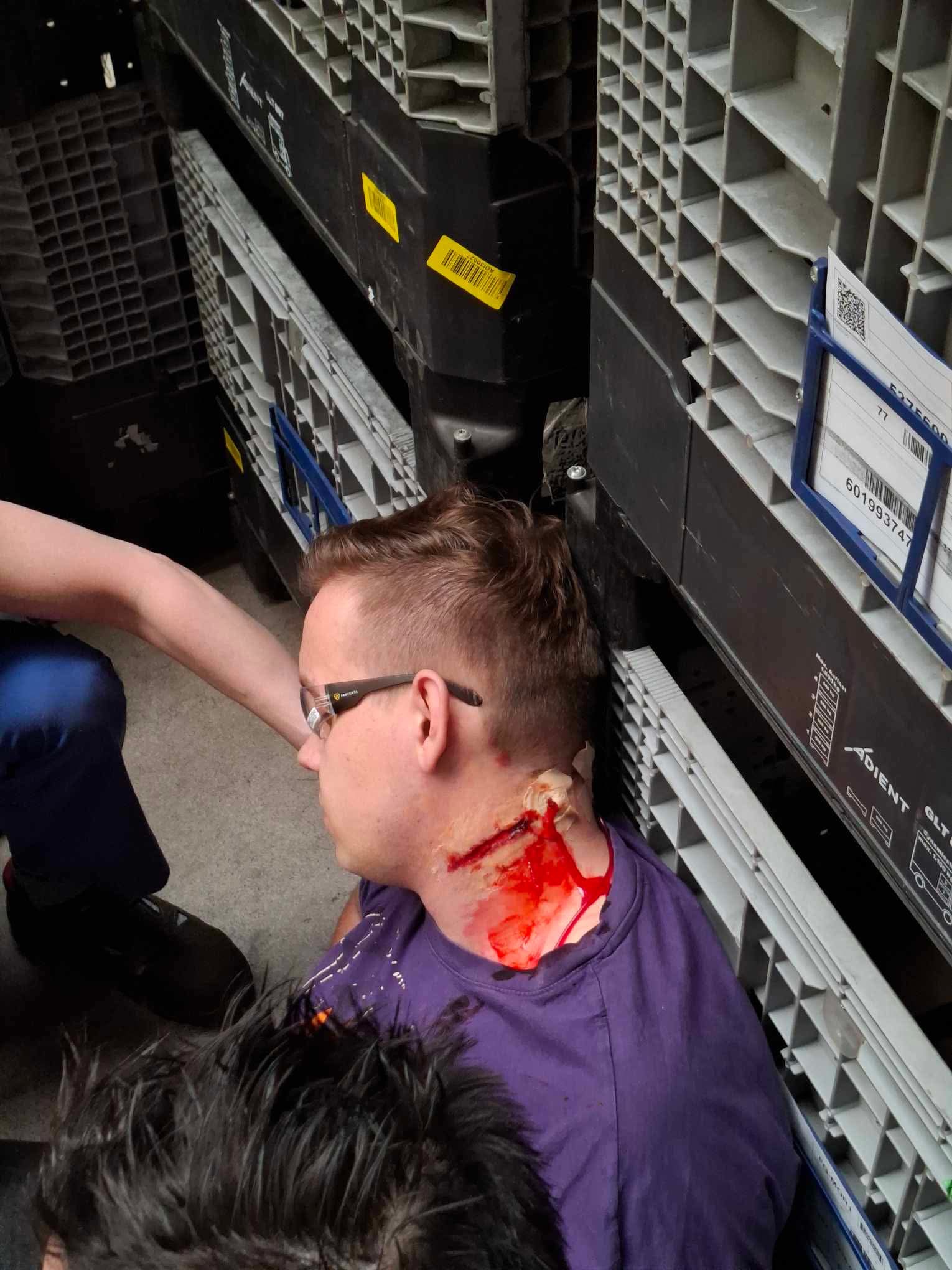 Symulacja rany szyi podczas kursu pierwszej pomocy. Mężczyzna z realistyczną raną, opatrywany przez instruktora w scenerii przypominającej magazyn.