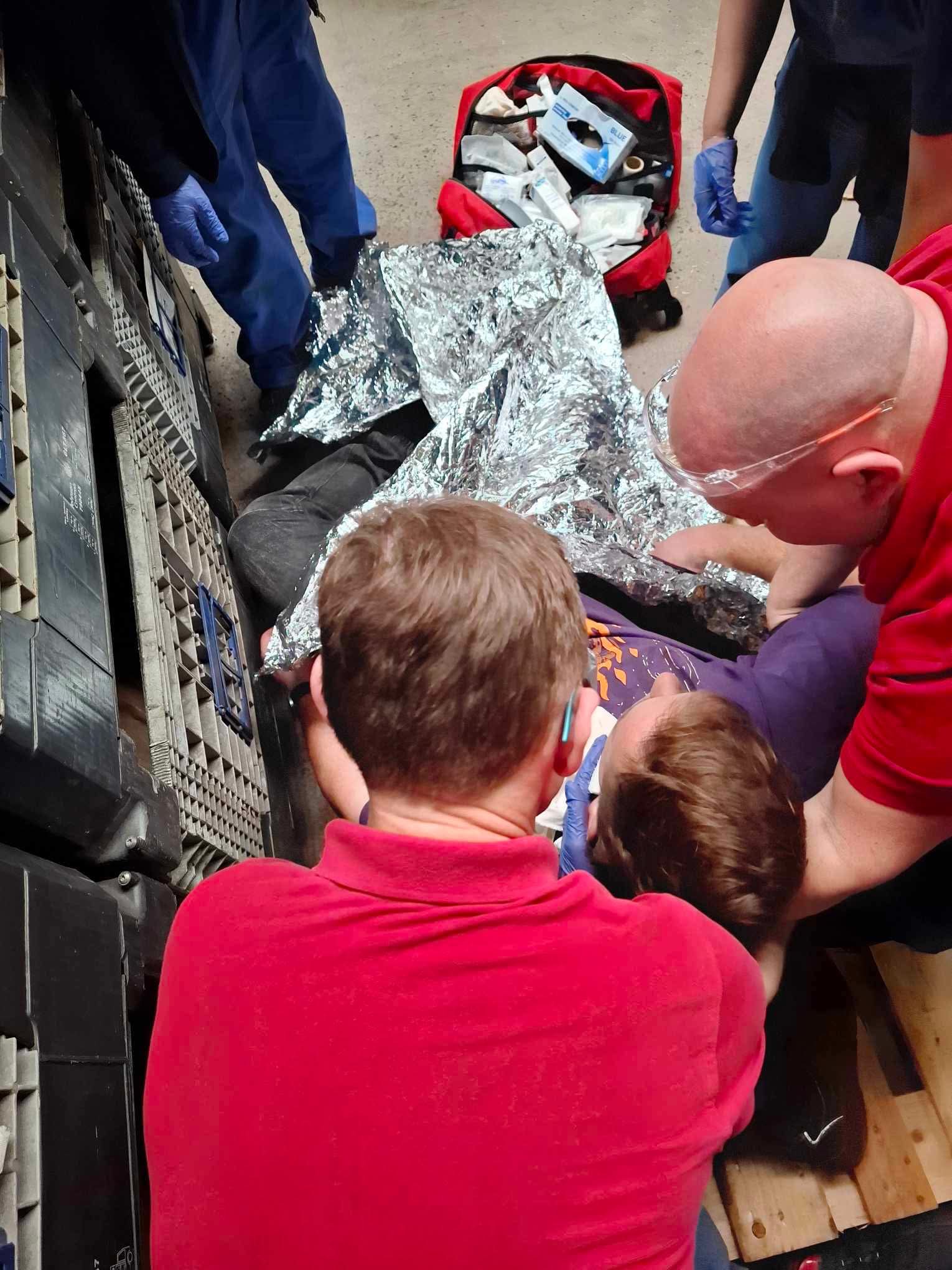 Symulacja udzielania pierwszej pomocy: mężczyzna leży na ziemi pod folią termiczną, otoczony przez osoby w rękawiczkach i czerwonych koszulkach. Widoczna apteczka.