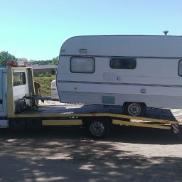 AUTO-POMOC.net - Biała przyczepa kempingowa zabezpieczona na platformie pojazdu transportowego z żółtymi elementami, widok z boku, w tle drzewa i budynek.