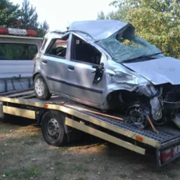 Srebrny samochód osobowy po wypadku, z poważnymi uszkodzeniami karoserii i rozbitą szybą przednią, transportowany na platformie pojazdu pomocy drogowej. Na dachu lawety widoczny pomarańczowy kogut.