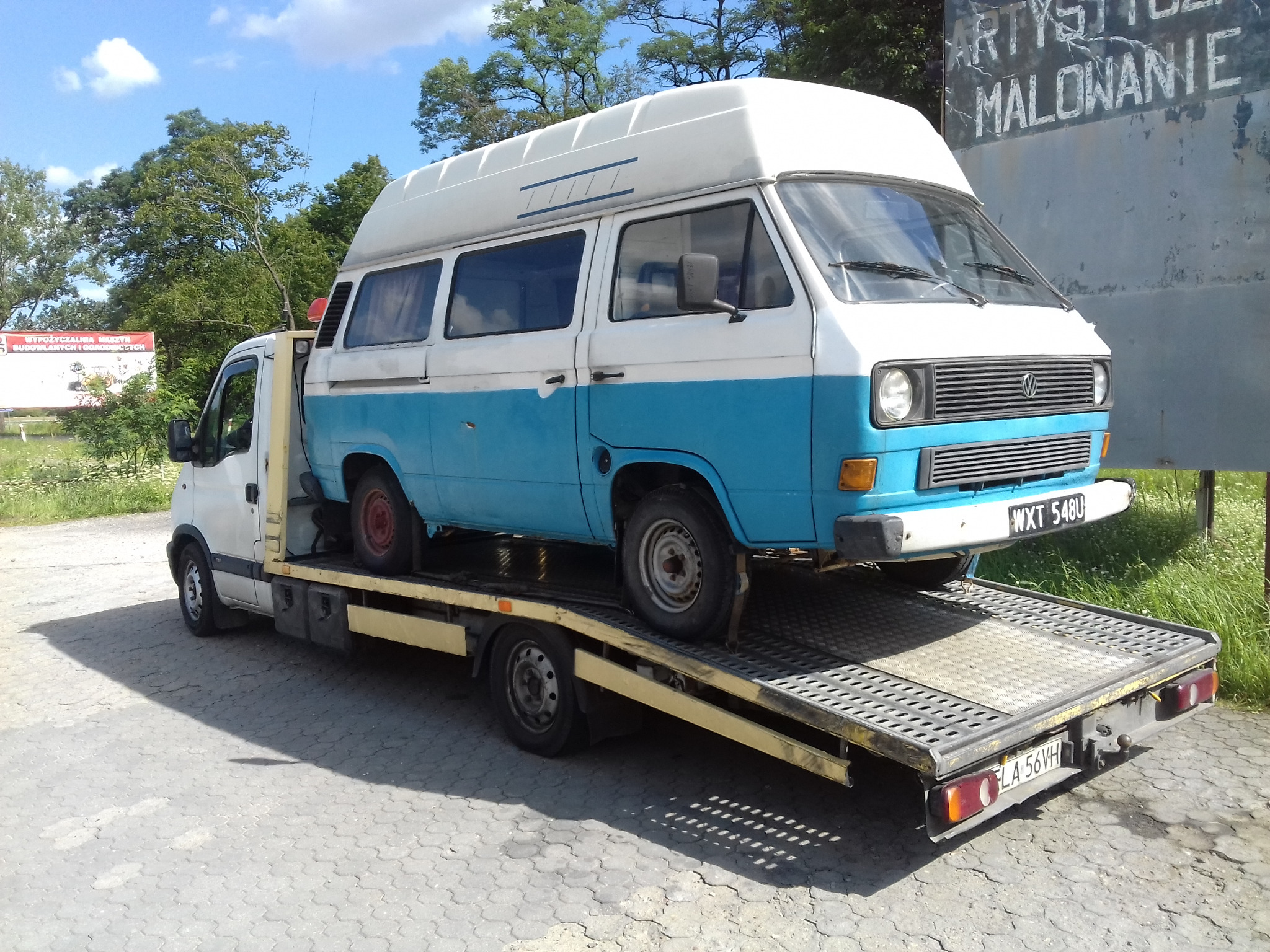 Biało-niebieski bus Volkswagen Transporter na platformie pojazdu pomocy drogowej, z tablicą rejestracyjną WXT 548U, w tle zieleń i tablica reklamowa.