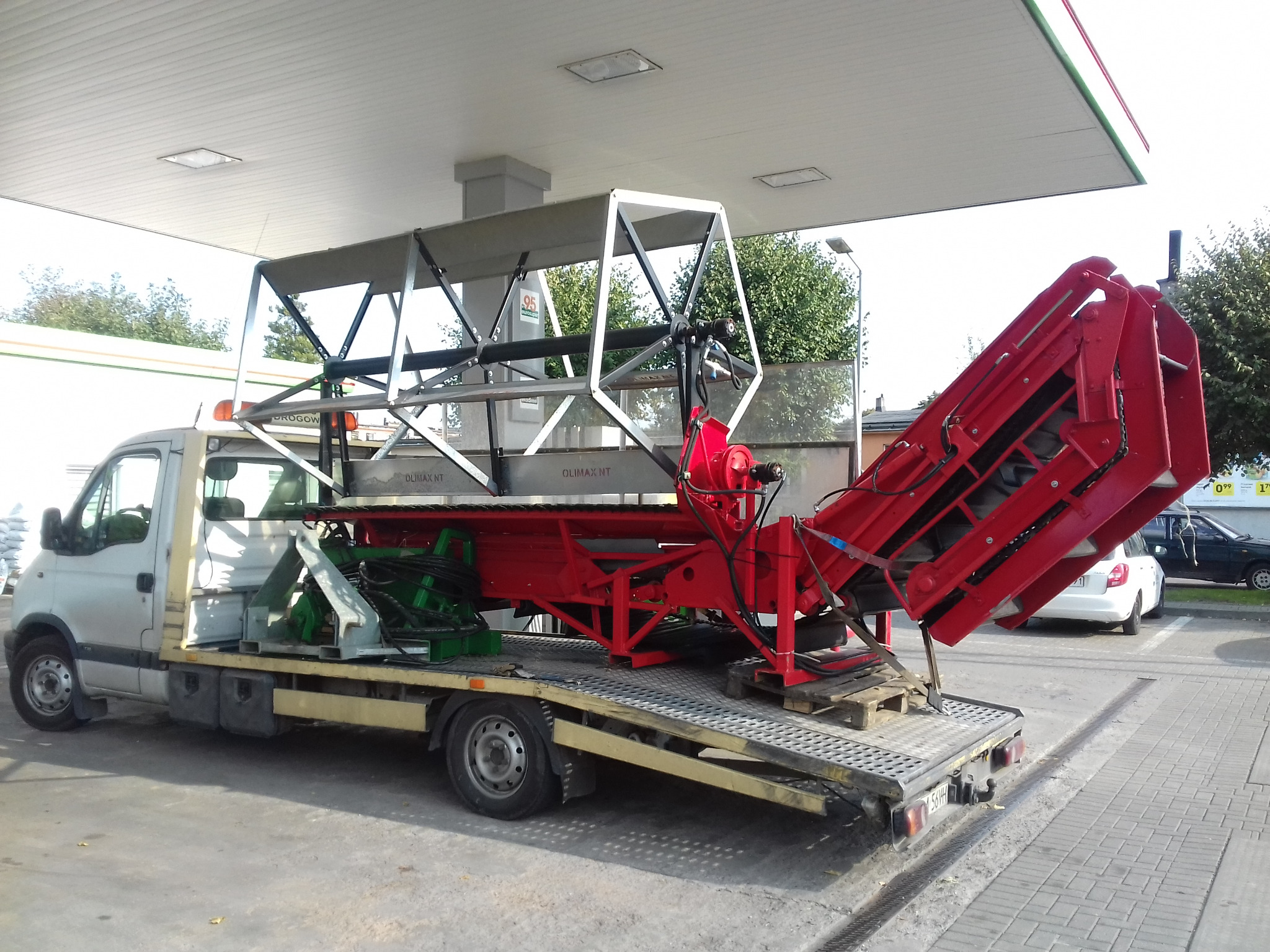 Biały samochód dostawczy z platformą transportową, przewożący czerwoną konstrukcję metalową z elementami hydraulicznymi, stojący na stacji benzynowej.