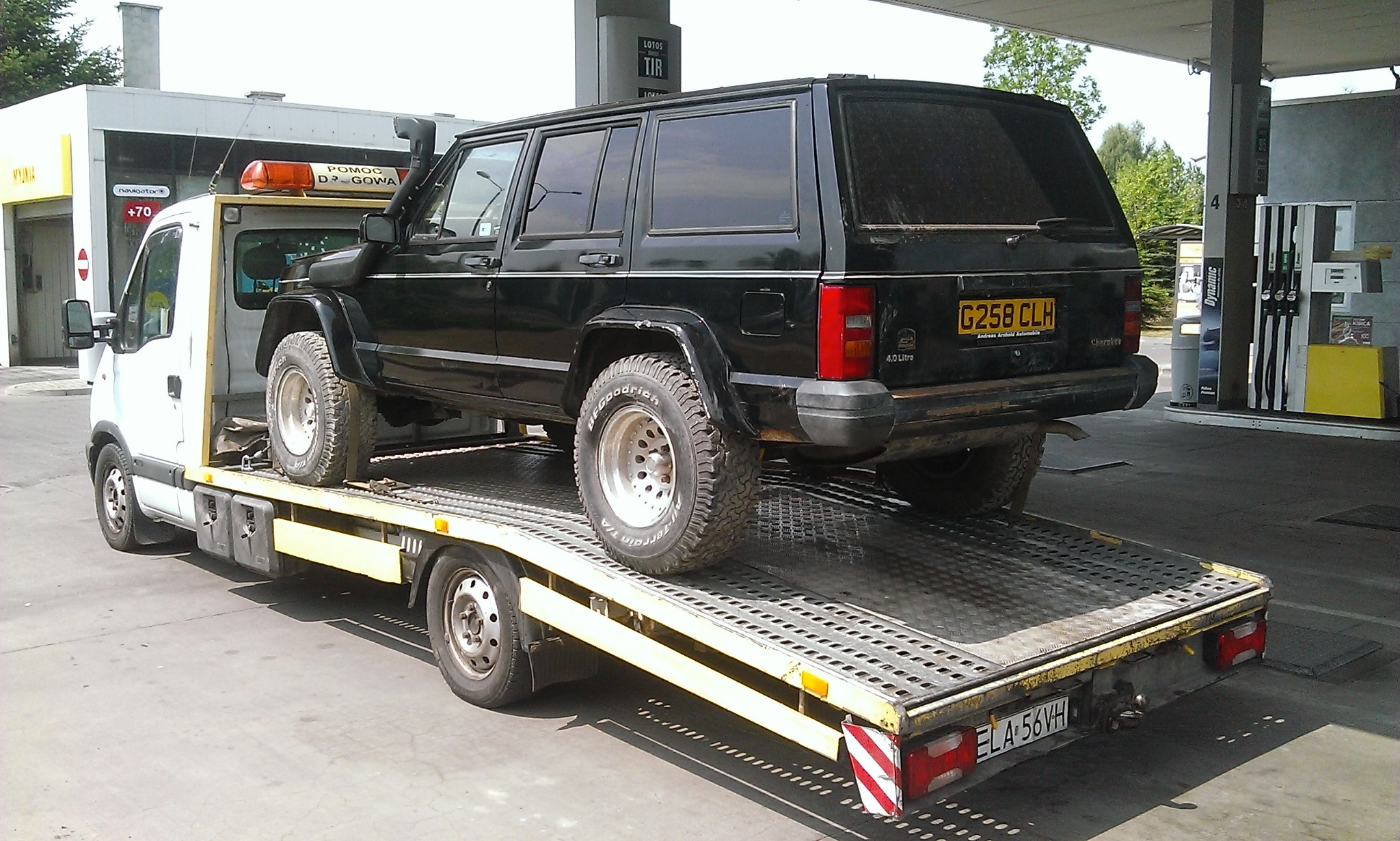 Czarny Jeep Cherokee 4.0 Litre na żółtej lawecie z lampą ostrzegawczą 'Pomoc Drogowa', na tle stacji benzynowej z dystrybutorami i myjnią.