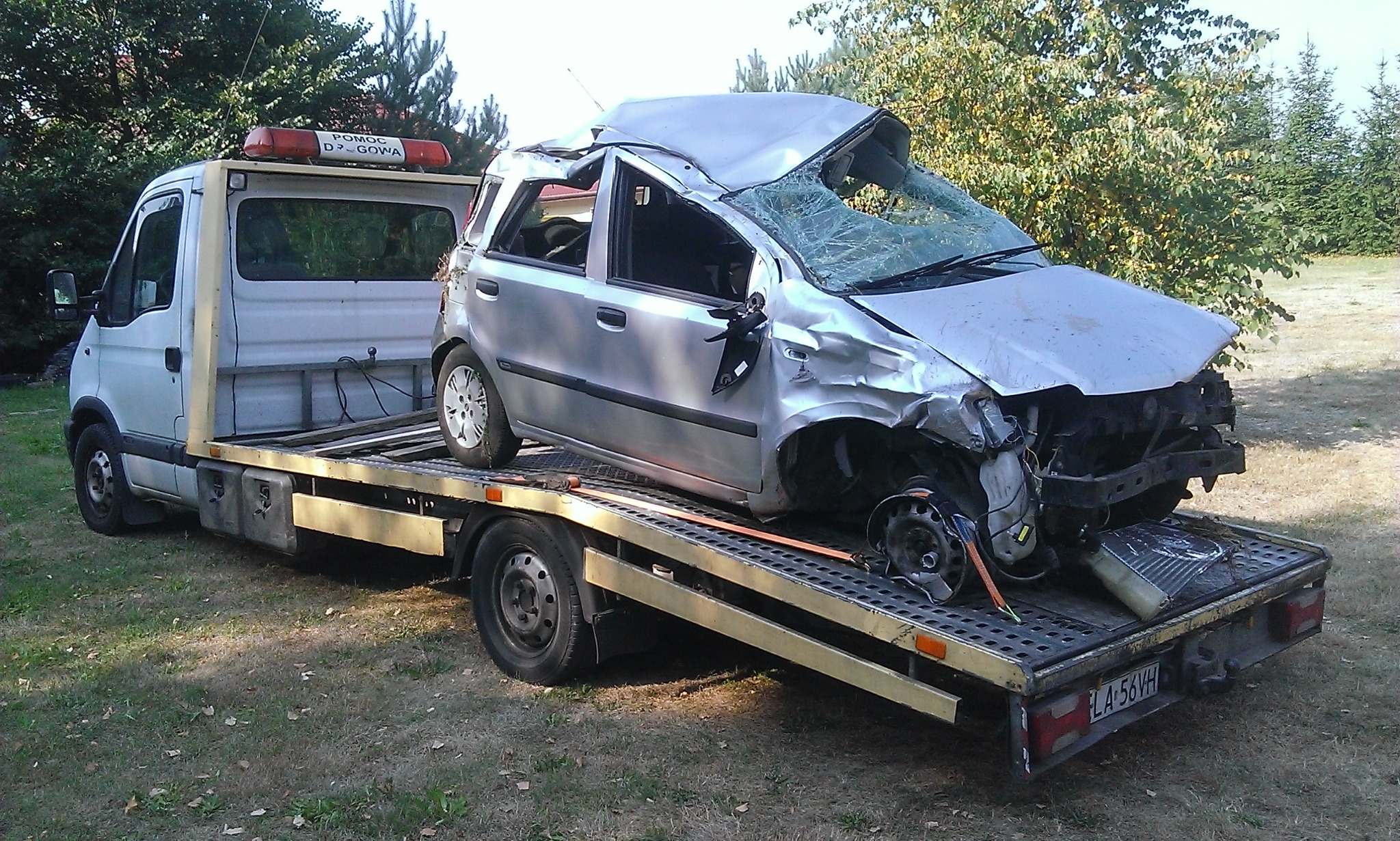 Srebrny samochód osobowy po wypadku, z poważnymi uszkodzeniami karoserii i rozbitą szybą przednią, transportowany na platformie pojazdu pomocy drogowej. Na dachu lawety widoczny pomarańczowy kogut.