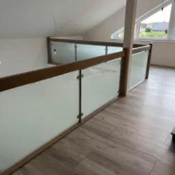 Wewnętrzna balustrada z drewnianymi elementami i matowym szkłem, zamontowana na antresoli w domu z poddaszem, drewniana podłoga, widok na okno dachowe i fragment ogrodu.