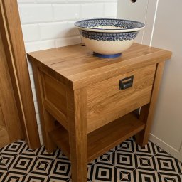 Mazurska Pracownia Drewna Dorota Kowalczyk - Stylowa szafka łazienkowa z drewna dębowego z szufladą i półką, na której stoi ceramiczna umywalka w niebieskie wzory, na tle białych płytek i podłogi w geometryczne wzory.