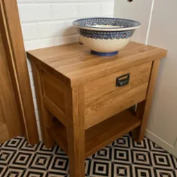 Stylowa szafka łazienkowa z drewna dębowego z szufladą i półką, na której stoi ceramiczna umywalka w niebieskie wzory, na tle białych płytek i podłogi w geometryczne wzory.