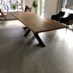 Mazurska Pracownia Drewna - Długi, dębowy blat stołu na czarnych, metalowych nogach w jasnym wnętrzu w Wygryny, z widokiem na okno i krzesła w tle.