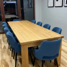 Duży, jasny stół z litego drewna w nowoczesnej jadalni, otoczony niebieskimi krzesłami z czarnymi nogami, podwieszane lampy ze szklanymi kloszami, parkiet z jasnego drewna, minimalistyczne obrazy...