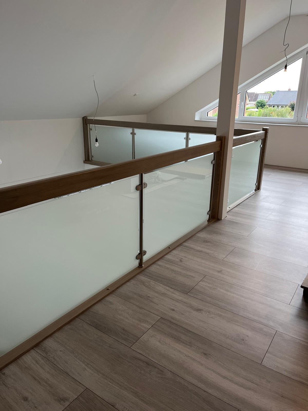 Wewnętrzna balustrada z drewnianymi elementami i matowym szkłem, zamontowana na antresoli w domu z poddaszem, drewniana podłoga, widok na okno dachowe i fragment ogrodu.