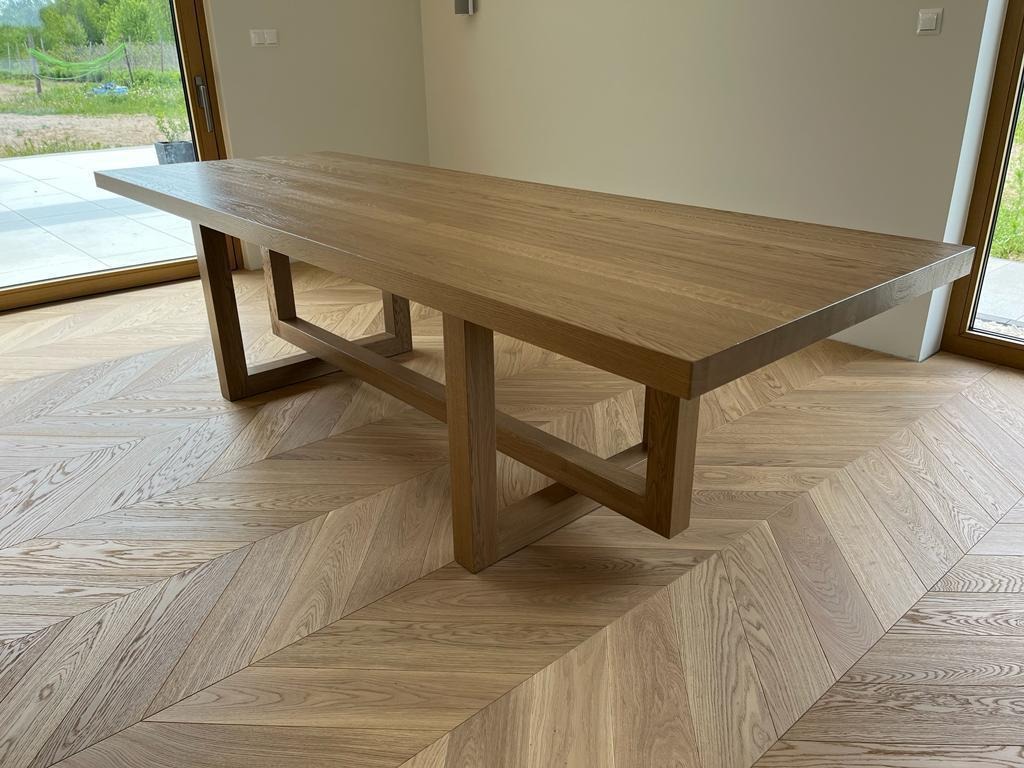 Stół z jasnego drewna o prostokątnym blacie i geometrycznych nogach, ustawiony na parkiecie w jodełkę, widok z perspektywy podłogi.