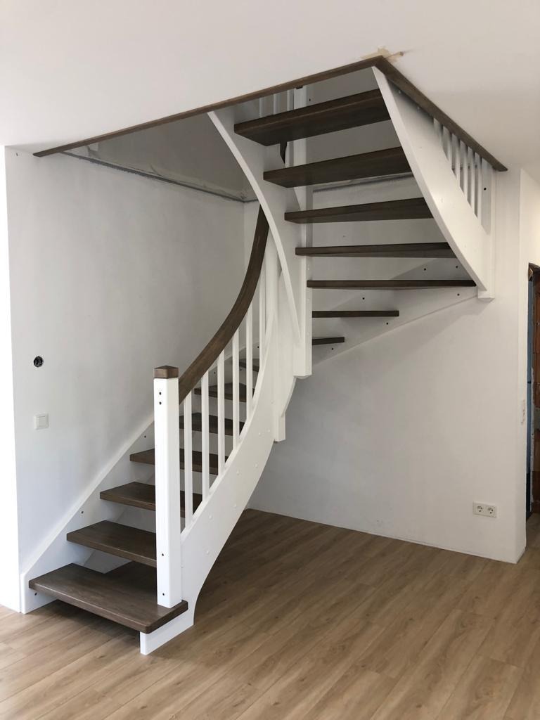 Kręte schody z drewnianymi stopniami i białą balustradą w jasnym wnętrzu, widoczne niedoskonałości wykończenia sufitu.