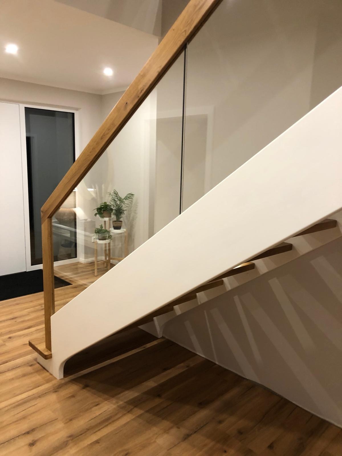 Nowoczesne schody z białymi stopniami i drewnianą poręczą z elementami szkła, widoczne rośliny w tle.