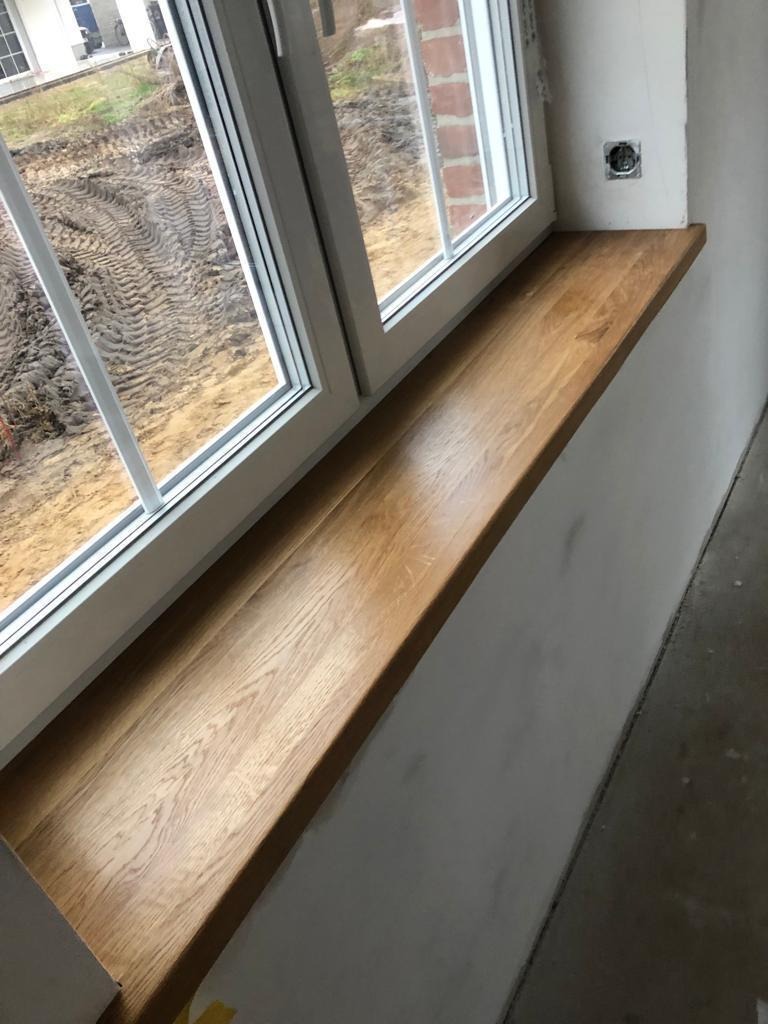 Jasny, dębowy parapet drewniany, zamontowany w białym oknie PCV, widok na plac budowy z ziemią i śladami opon.