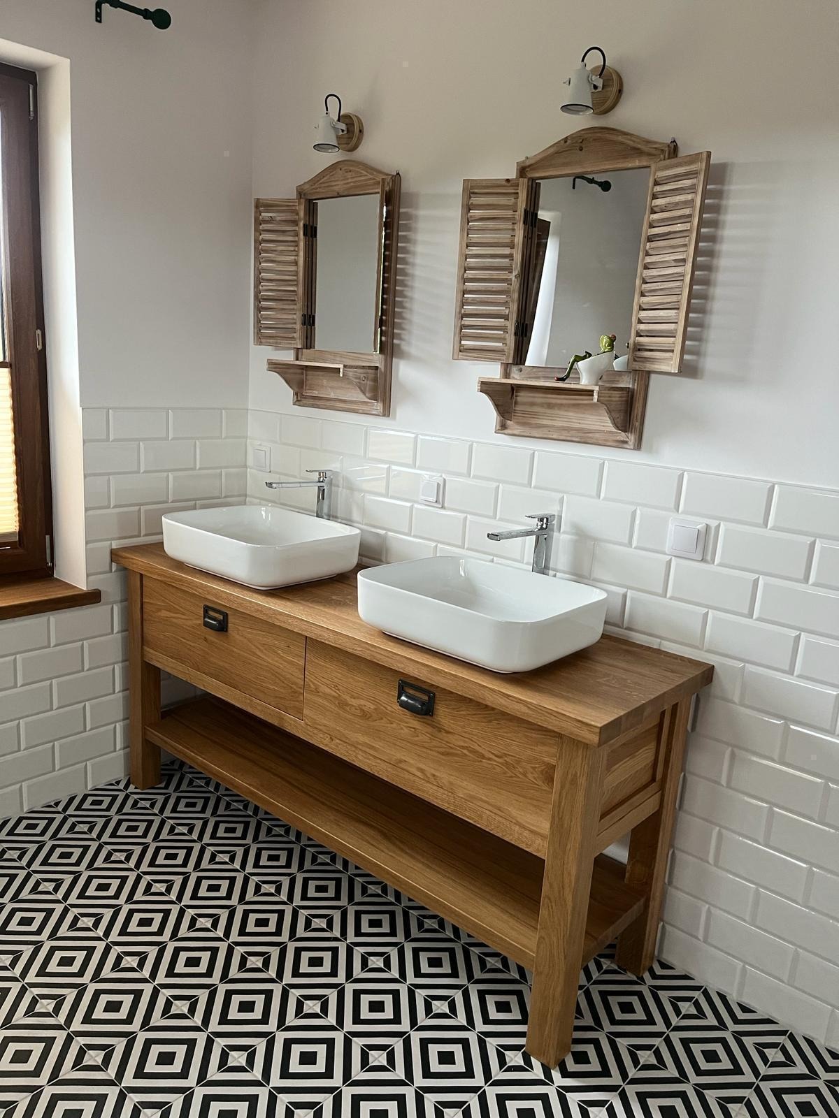 Łazienka z dwiema białymi umywalkami nablatowymi na drewnianej szafce z szufladami i półką, nad którymi wiszą lustra w drewnianych ramach z okiennicami, białe płytki na ścianach i wzorzysta...