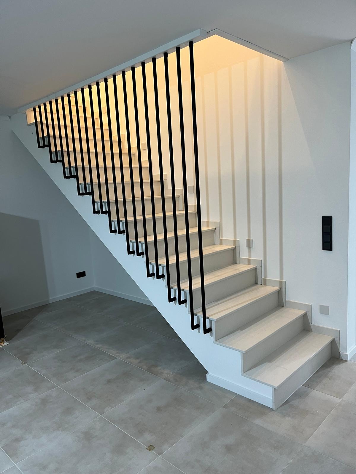 Nowoczesne schody o białych stopniach i czarnych, minimalistycznych balustradach, oświetlone od góry, z szarymi płytkami podłogowymi.