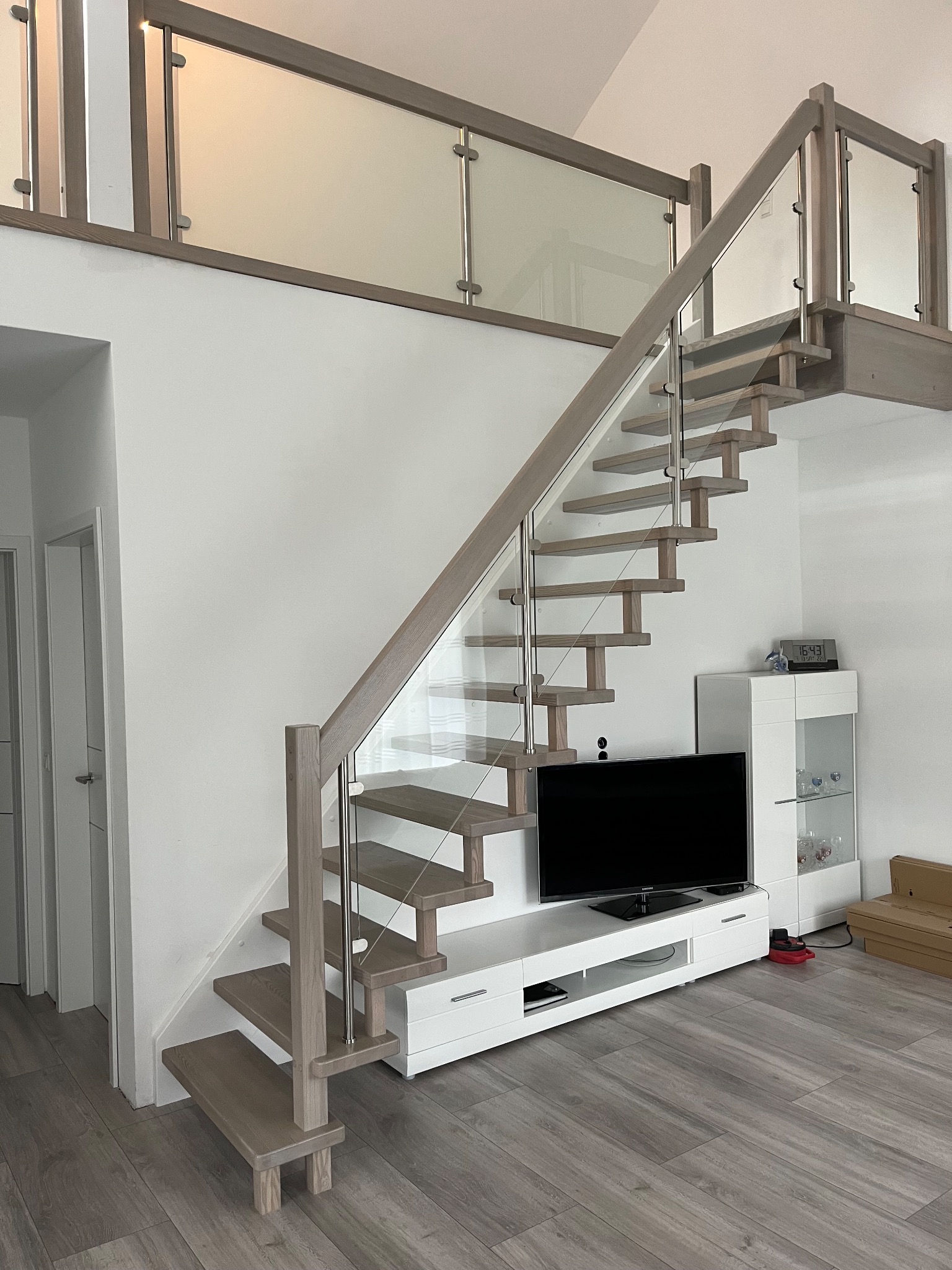 Nowoczesne schody z drewnianymi stopniami i szklaną balustradą, prowadzące na antresolę z podobną szklaną balustradą. W tle biała ściana i meble w salonie.