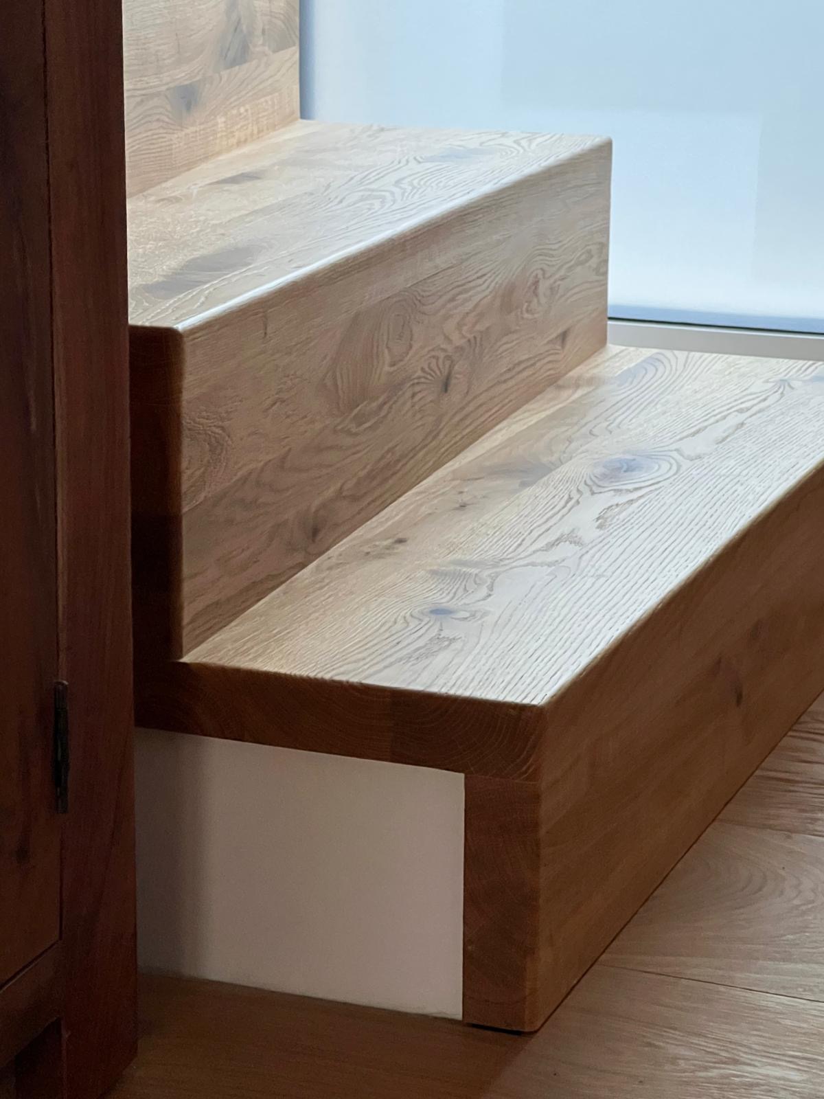 Dwa stopnie drewnianych schodów z jasnego drewna dębowego, z widocznym usłojeniem, osadzone na białej podstawie, przylegające do ciemnego drewnianego mebla i białej ściany.