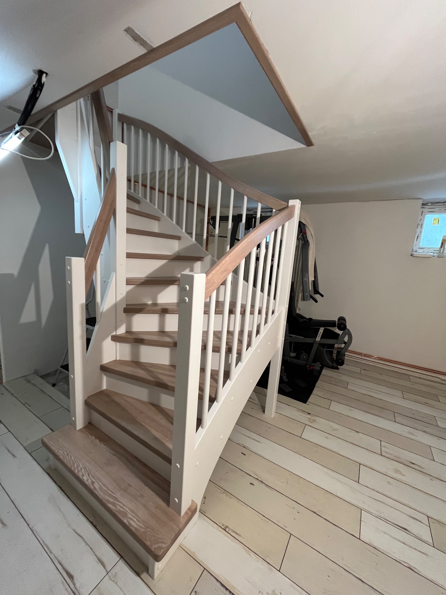 Widok z dołu na białe schody z drewnianymi stopniami i poręczą, prowadzące na wyższą kondygnację, z widocznym fragmentem sufitu i podłogi imitującej drewno, w tle sprzęt do ćwiczeń.