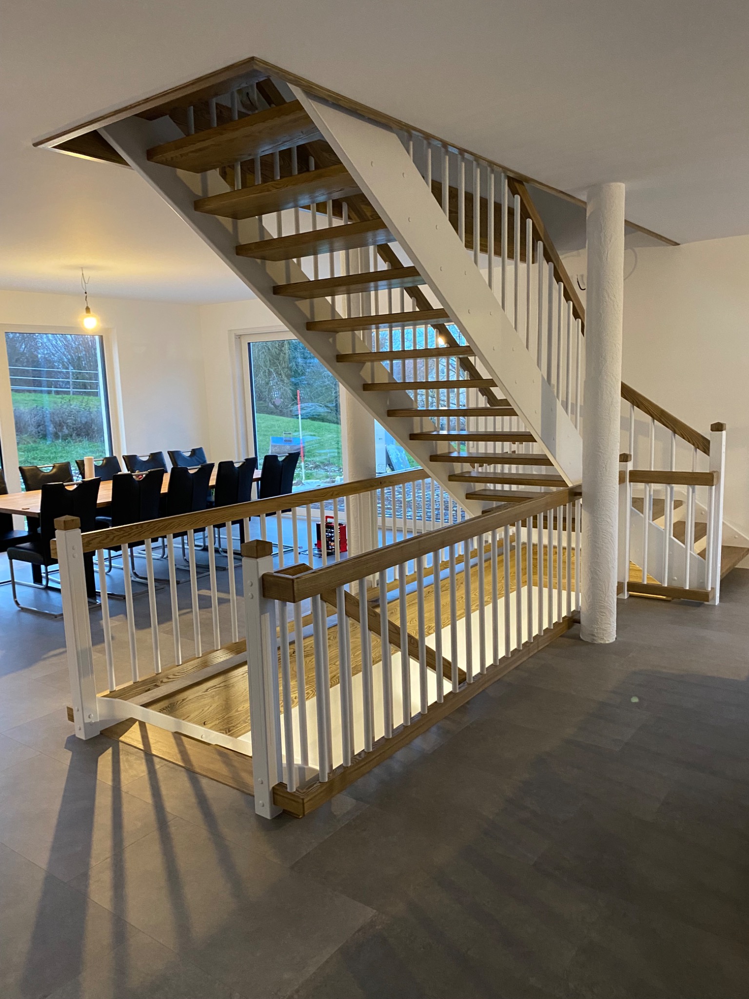 Jasne, drewniane schody z białymi balustradami w nowoczesnym wnętrzu domu; widok z dołu na górę, z widoczną jadalnią w tle.