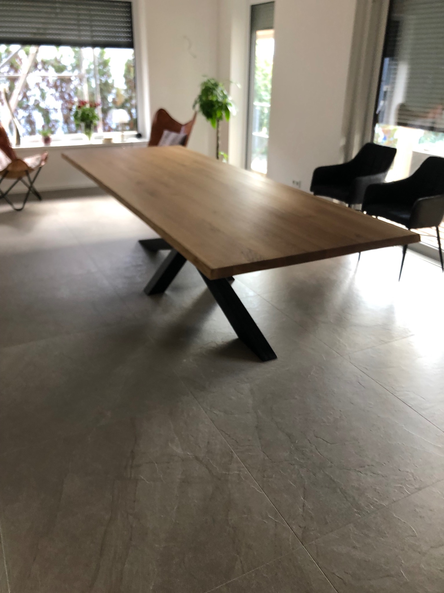 Długi, dębowy blat stołu na czarnych, metalowych nogach w jasnym wnętrzu w Wygryny, z widokiem na okno i krzesła w tle.