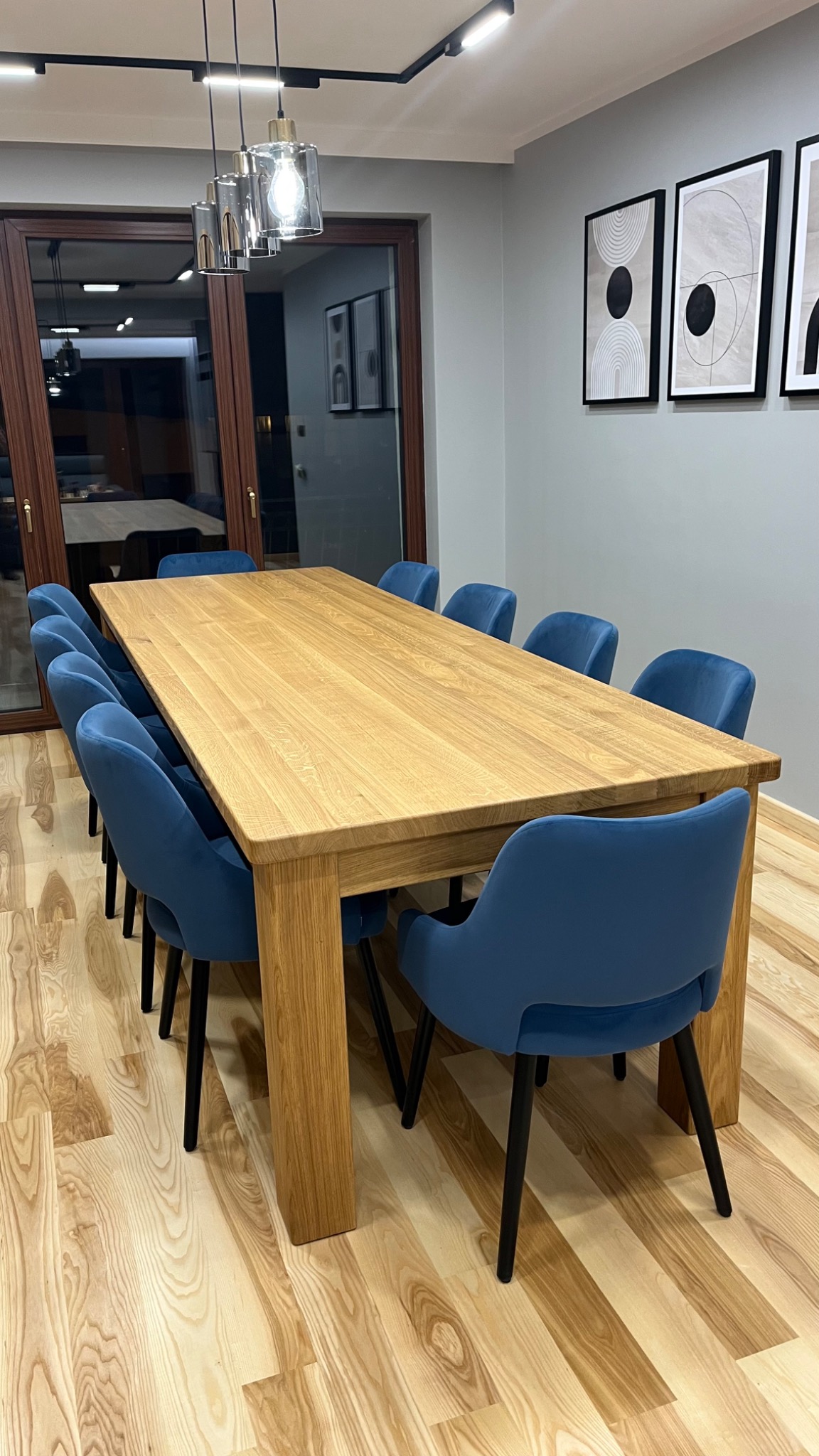 Duży, jasny stół z litego drewna w nowoczesnej jadalni, otoczony niebieskimi krzesłami z czarnymi nogami, podwieszane lampy ze szklanymi kloszami, parkiet z jasnego drewna, minimalistyczne obrazy...