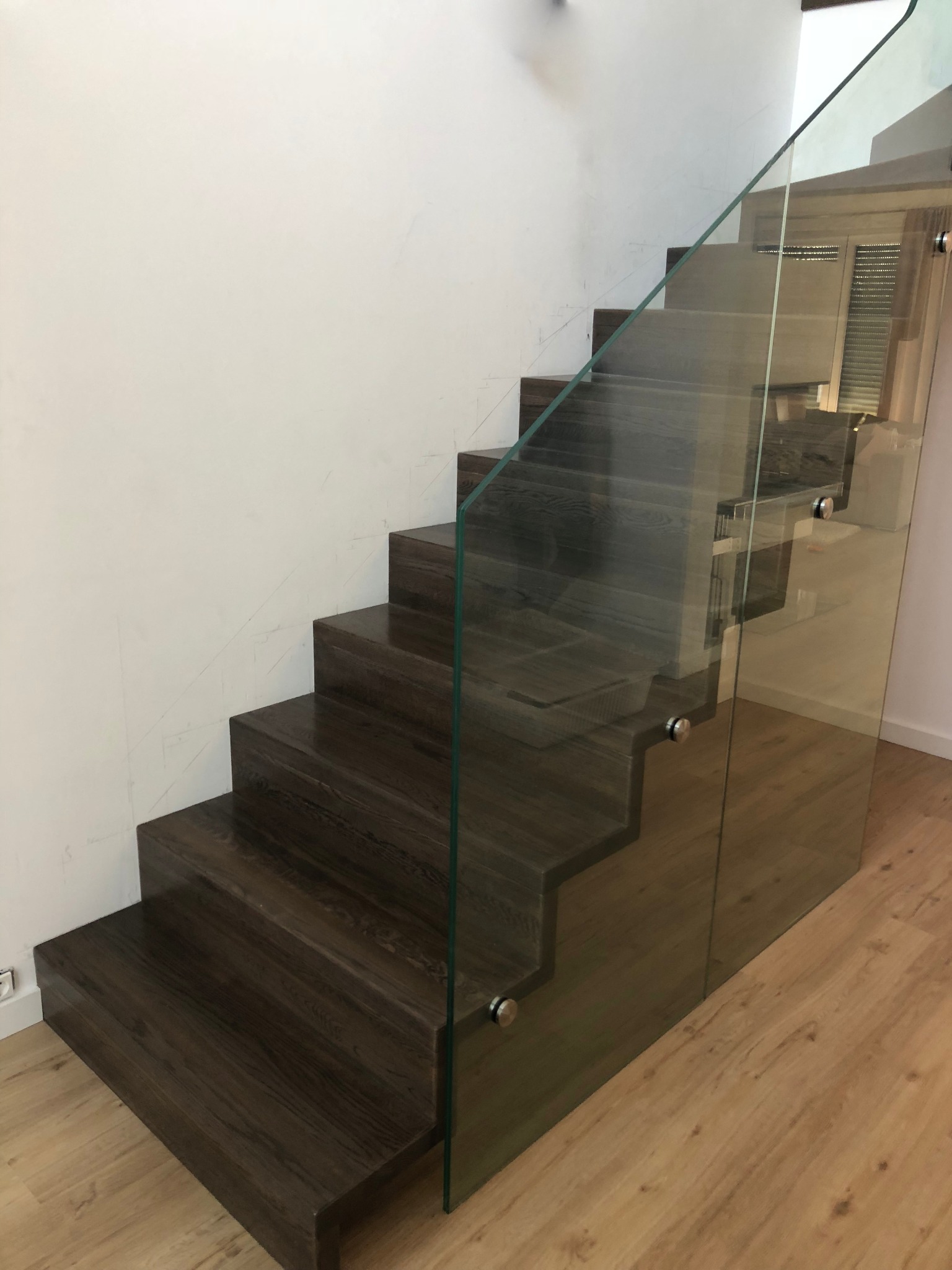 Nowoczesne schody z ciemnego drewna i szklaną balustradą, mocowaną na metalowych elementach, na tle jasnej ściany i podłogi.