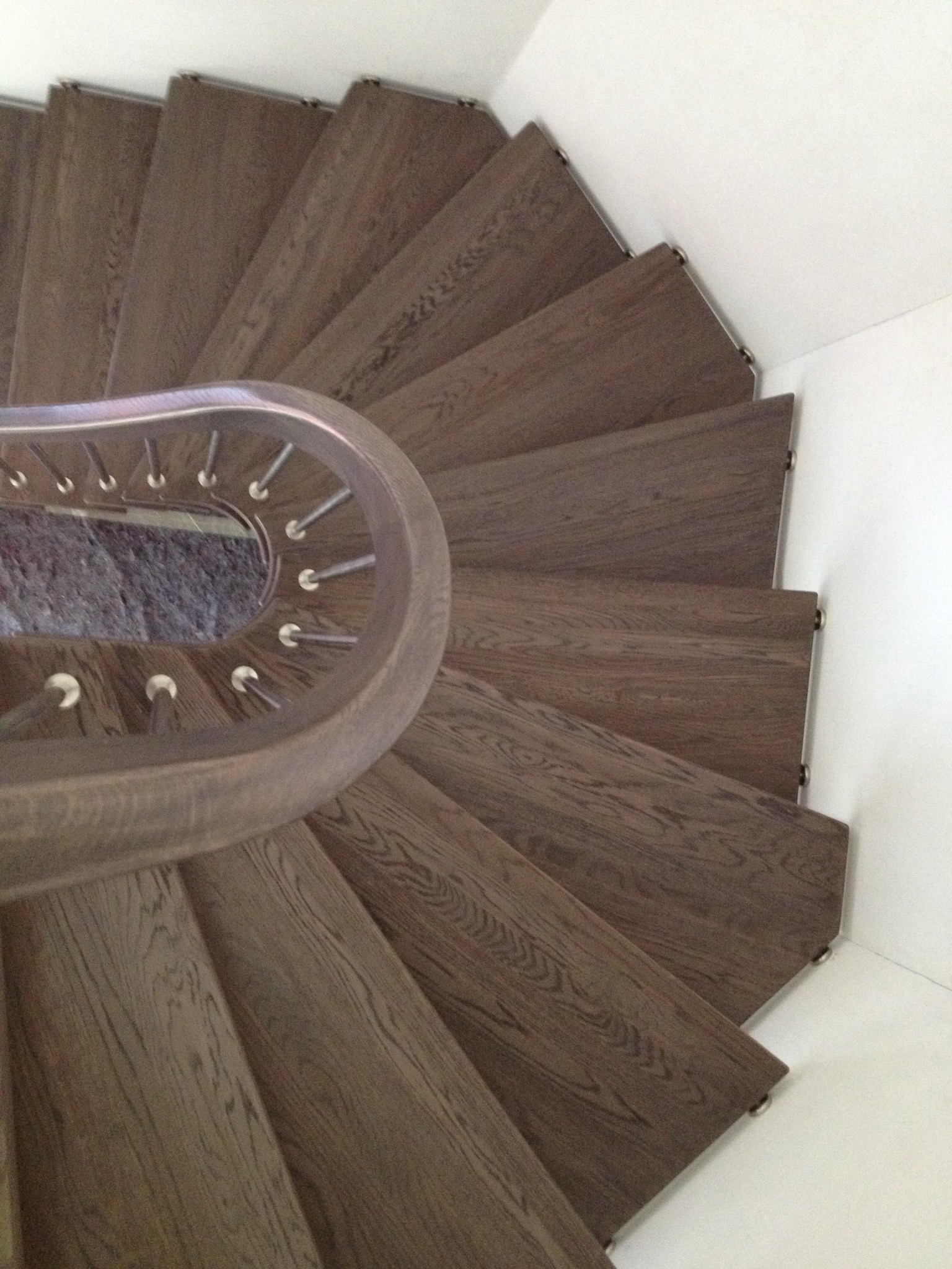 Spiralne schody drewniane z metalową poręczą, widok z góry. Ciemne drewno stopni kontrastuje z bielą ścian. Minimalistyczny design.