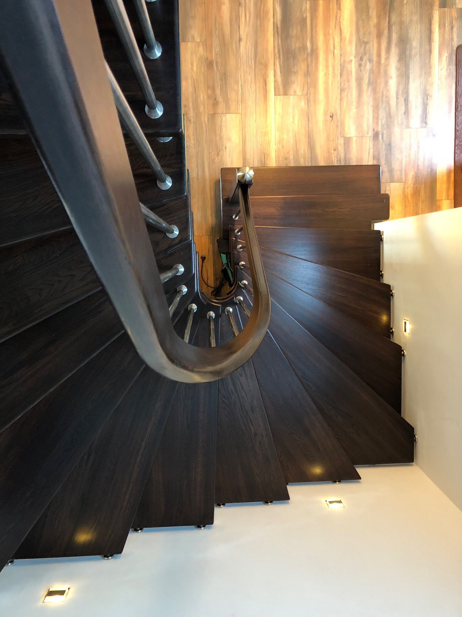 Spiralne schody z ciemnego drewna z metalową poręczą, oświetlone punktowymi lampami podłogowymi i ściennymi. Widok z góry podkreśla kręty kształt i nowoczesny design.