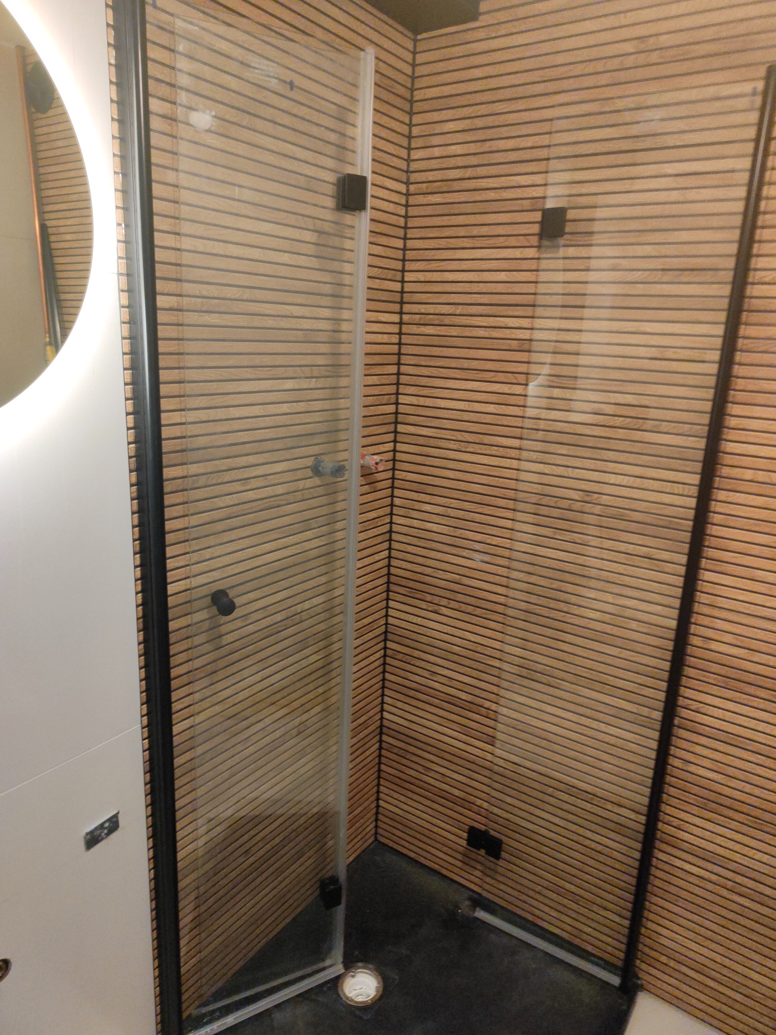 Nowoczesna kabina prysznicowa z czarnymi profilami i drewnianą okładziną ścienną. Okrągłe lustro z podświetleniem LED. Minimalistyczny design.