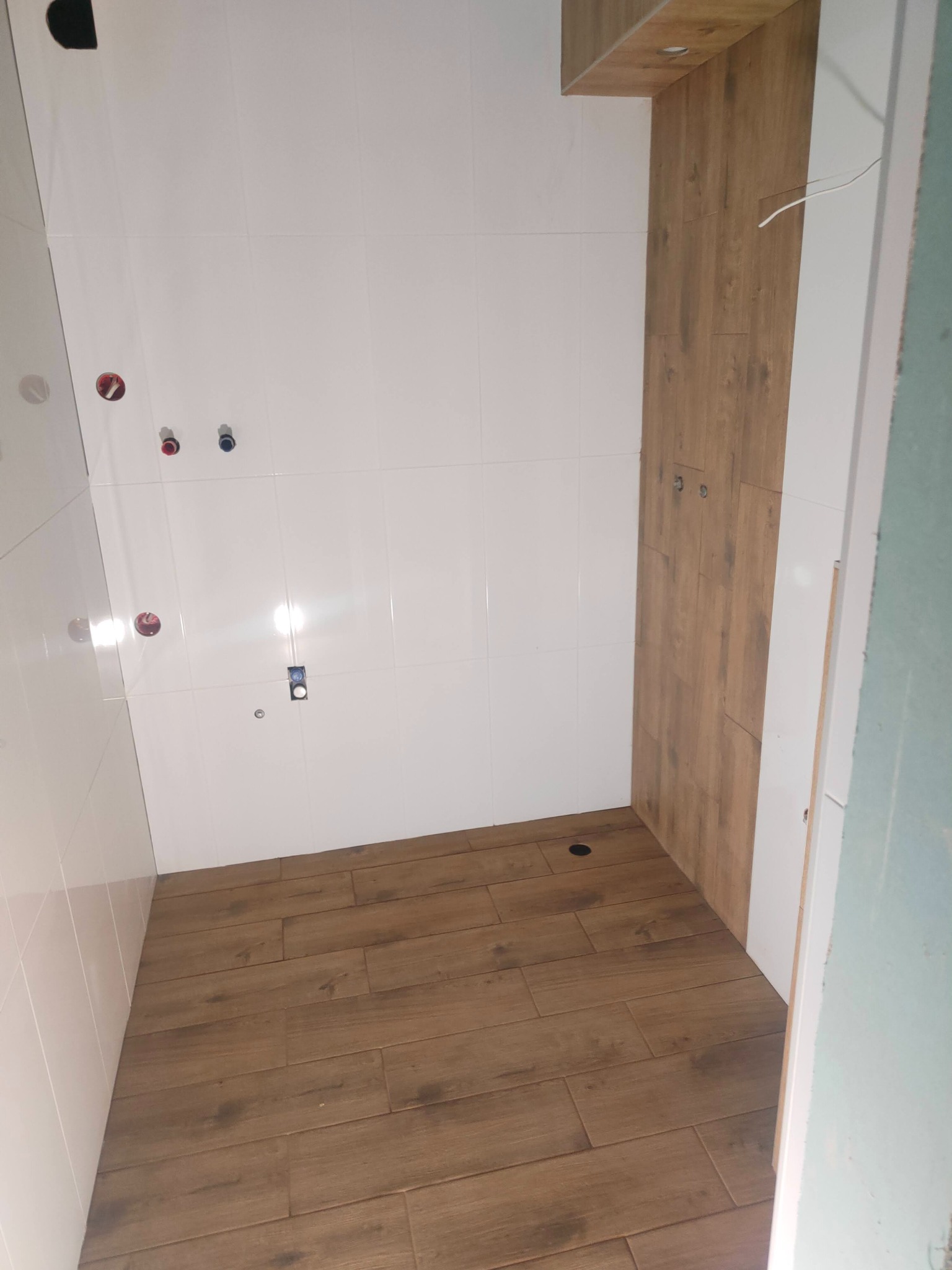 Wykończona łazienka z białymi płytkami ściennymi i podłogą imitującą drewno. Widoczne przyłącza wodne i elektryczne. Częściowo wykończona ściana z drewnopodobnych paneli.